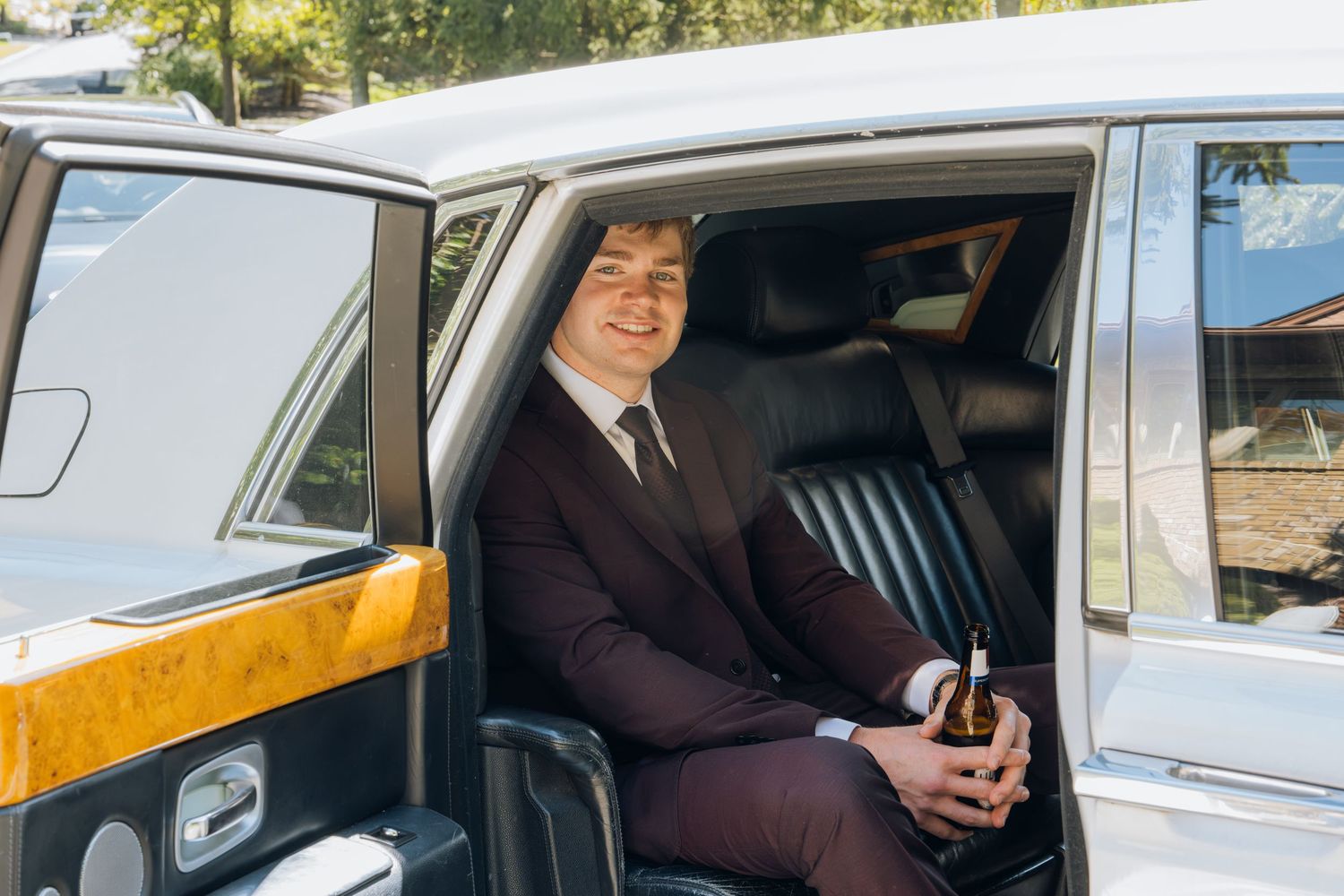 Smiling man in burgundy suit holding beer bottle seated in luxury white Rolls-Royce car backseat.