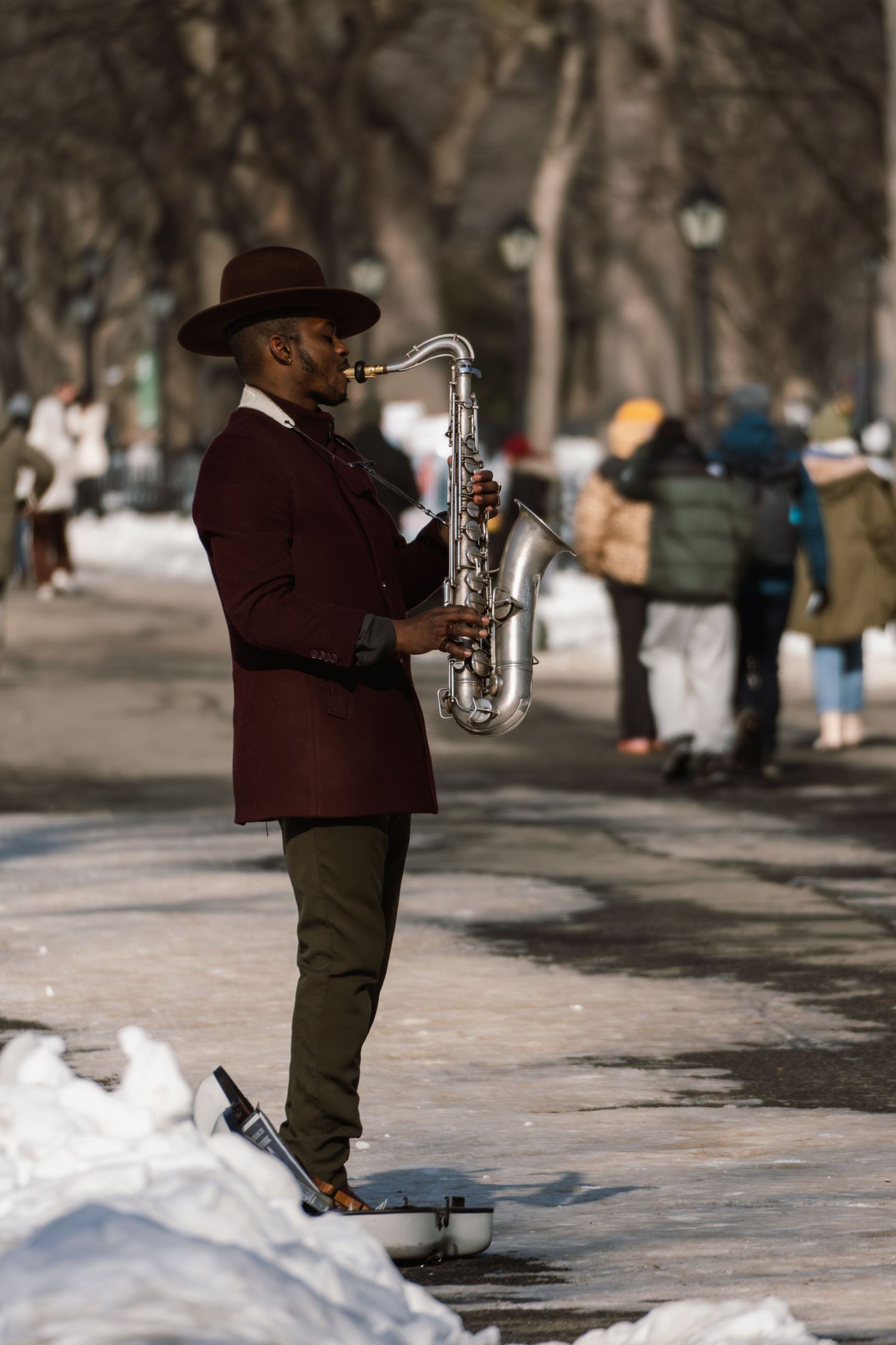 A stylish man in a burgundy jacket and wide-brim hat plays saxophone on a snowy park path.