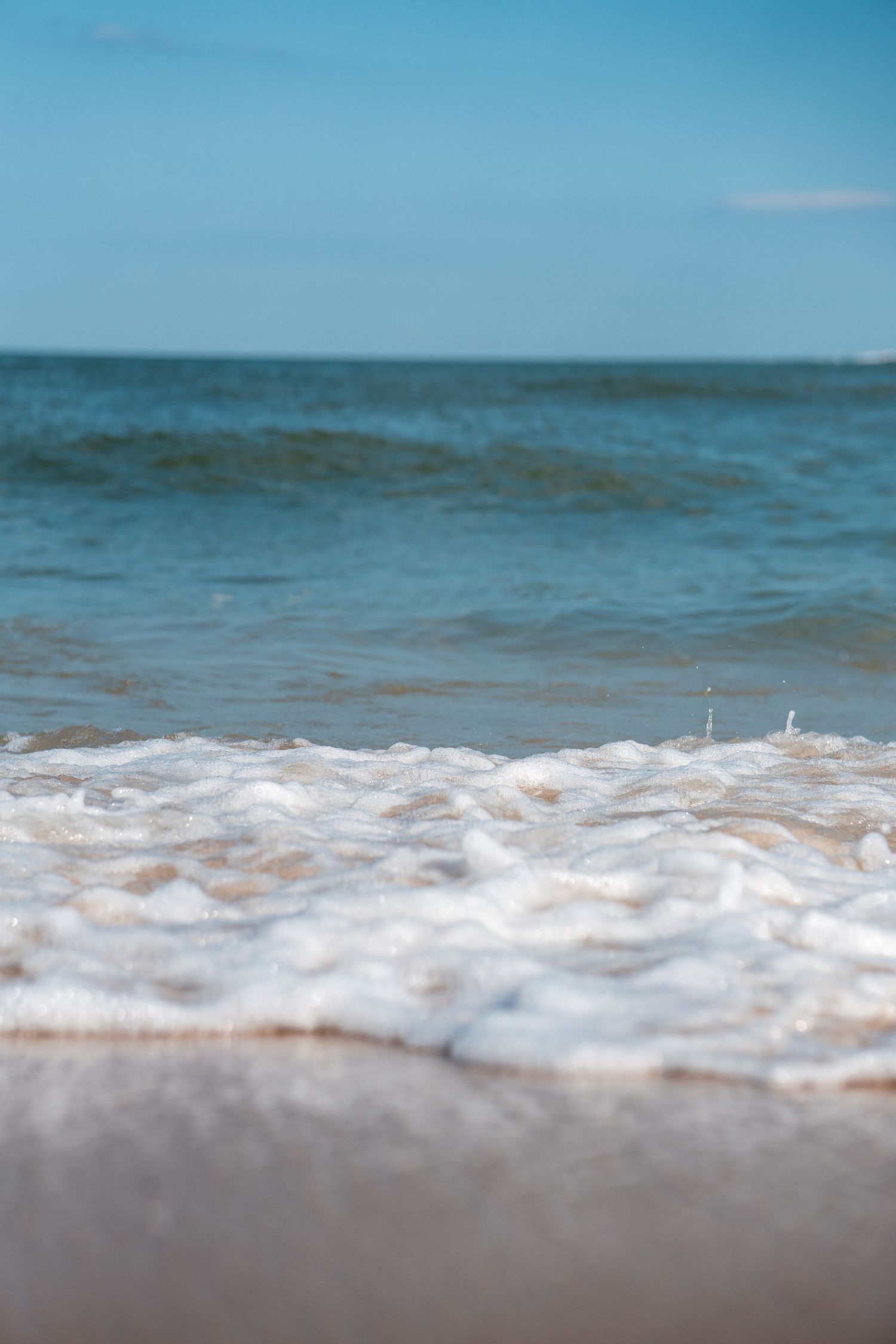 Close-up of white foamy ocean waves washing onto a sandy beach with calm blue water horizon.