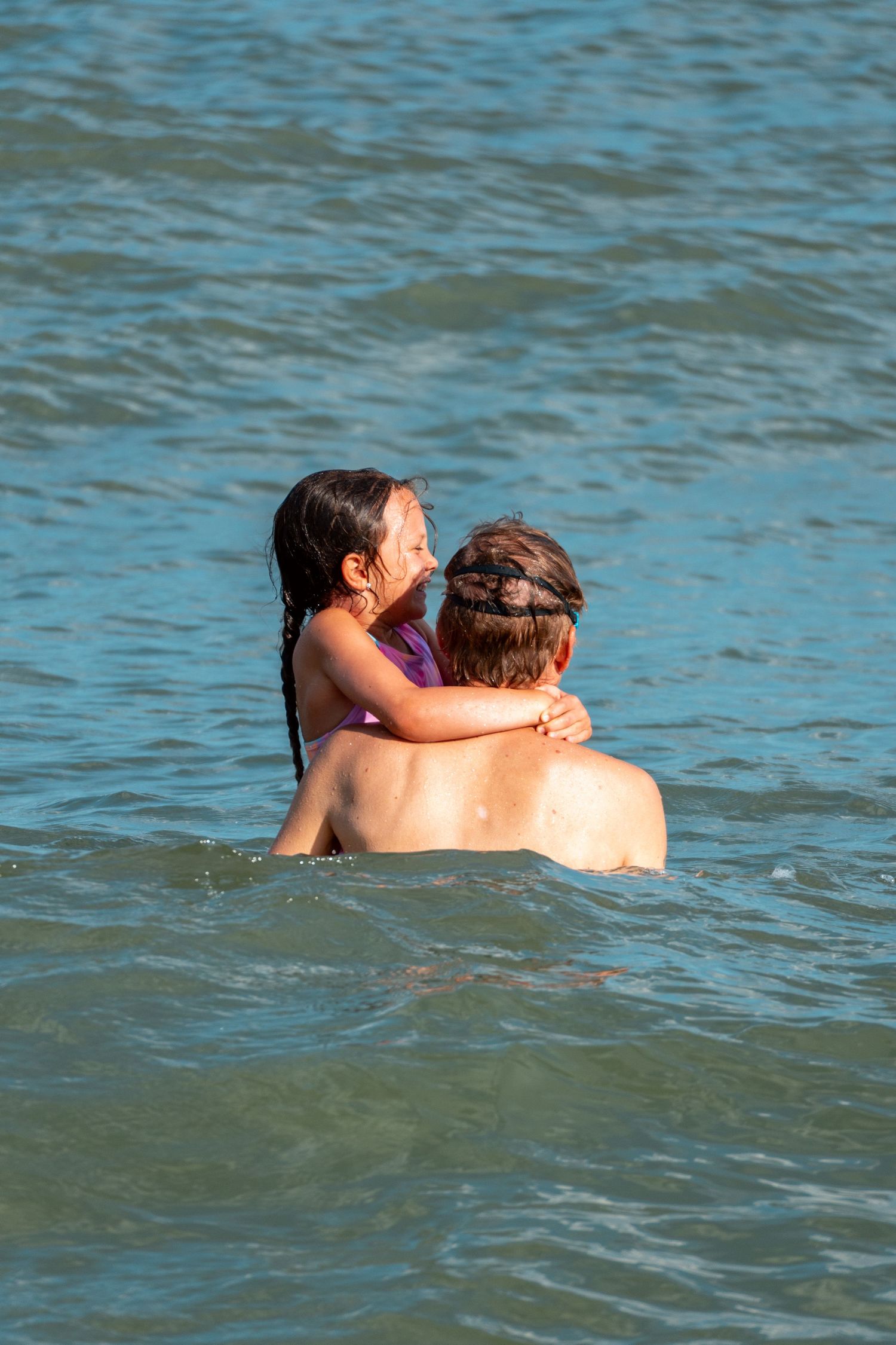 Young girl with braids hugging an adult from behind while swimming together in clear blue water.