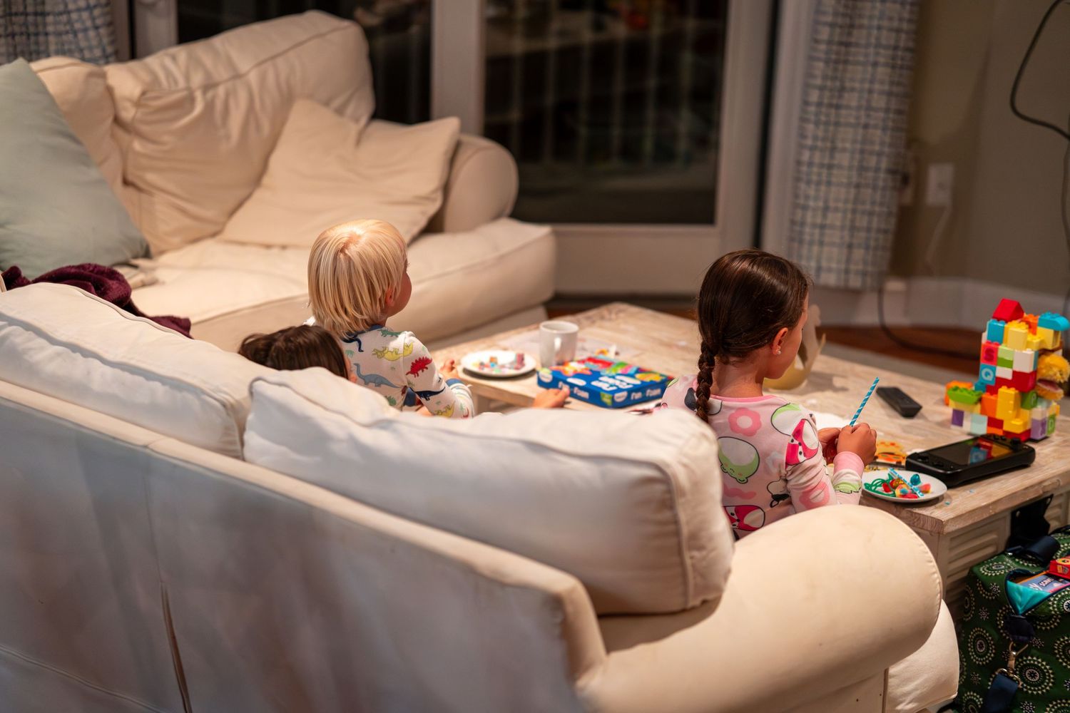 Two young children sitting behind a white sofa with snacks and toys on a table.