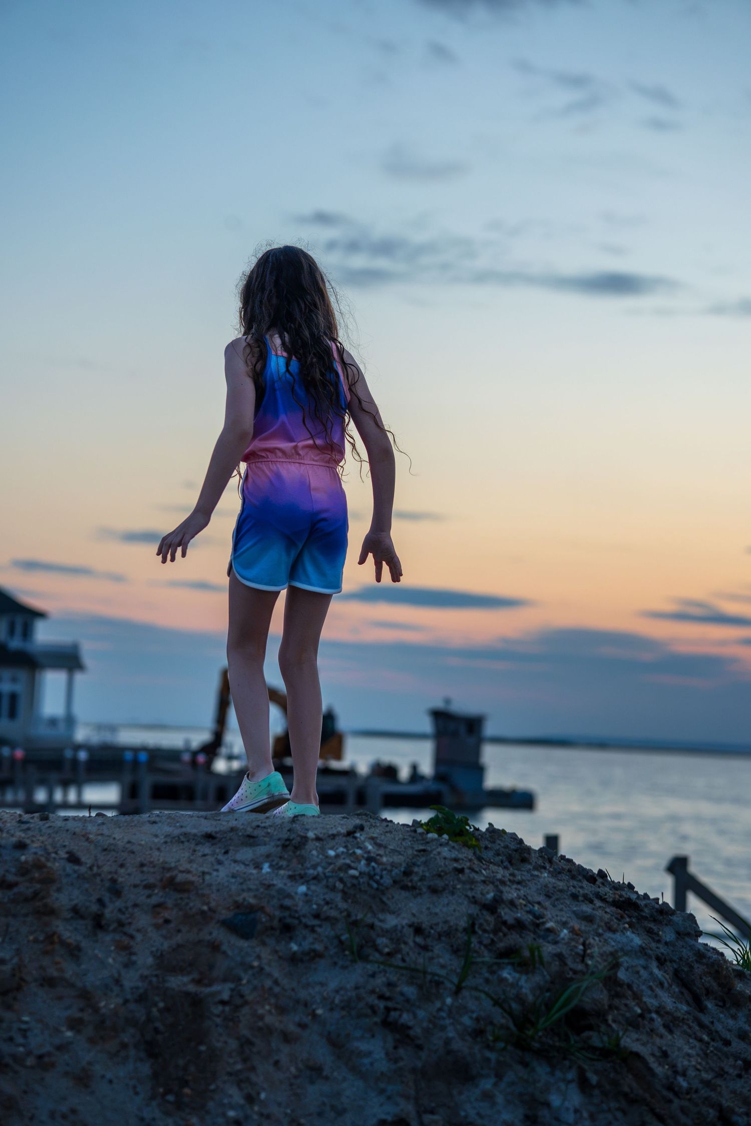 Girl in colorful romper stands on sandy bluff overlooking ocean pier at sunset.