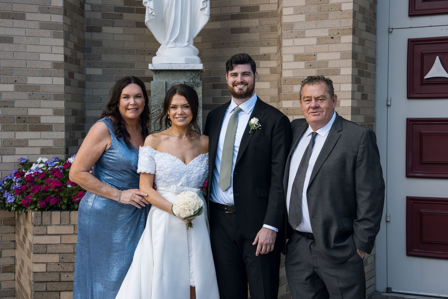 Wedding party of four posing outside a brick church, bride in white gown holding bouquet with family in formal attire.