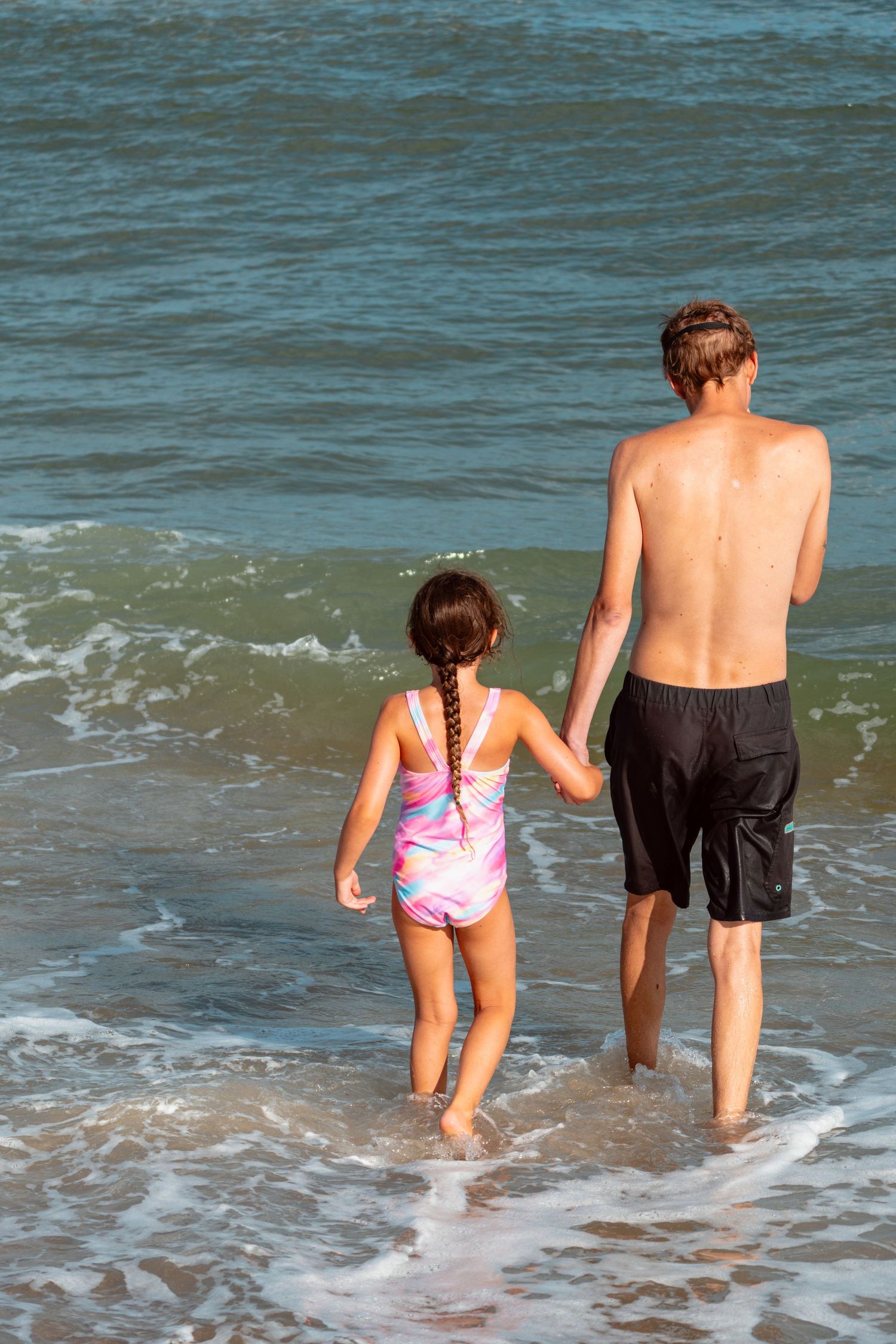 A teenager and young girl in swimwear hold hands walking into ocean waves from behind.