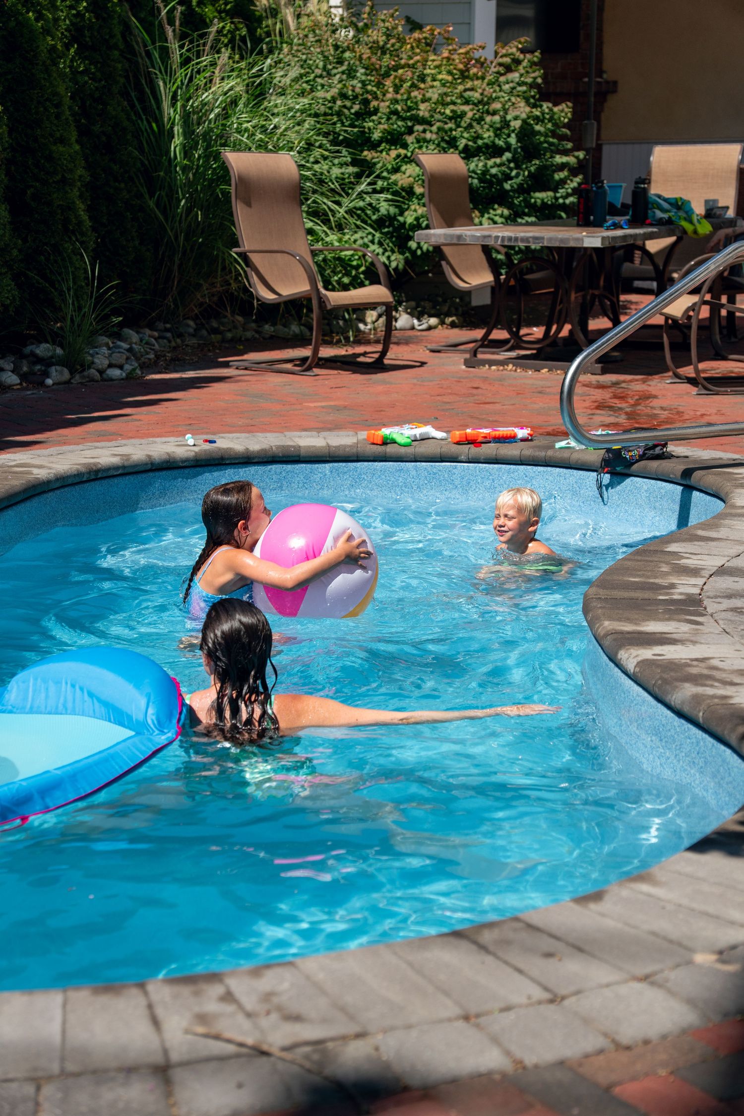Children playing with a pink beach ball in a backyard swimming pool on a sunny summer day.