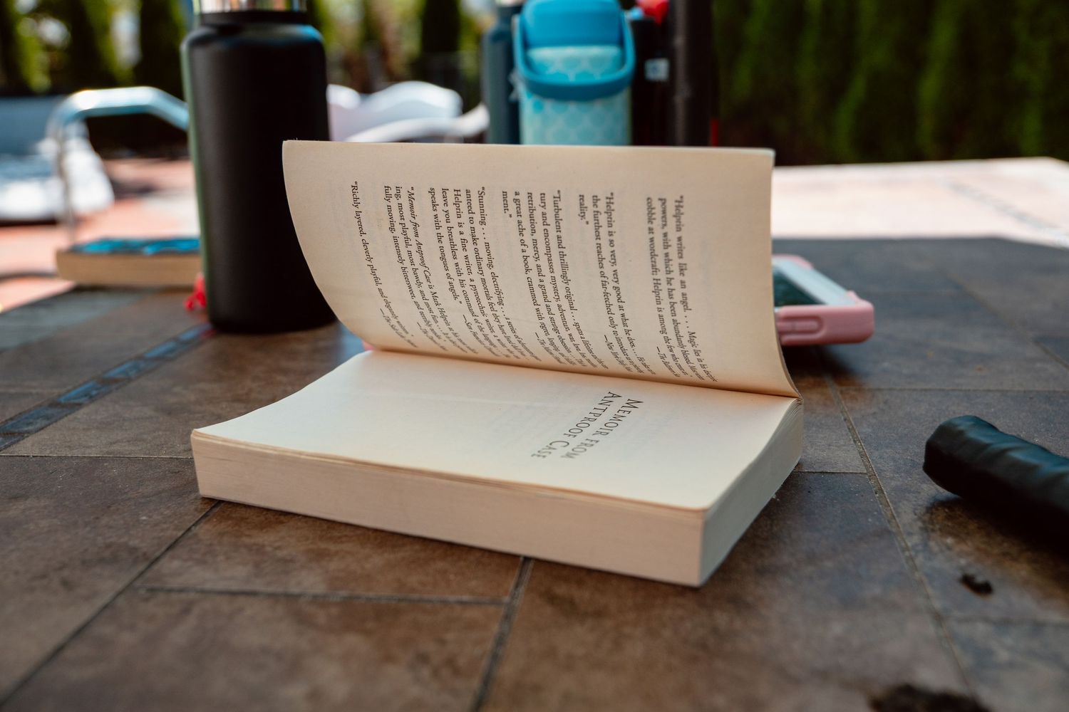 Open paperback book lying on a tiled outdoor table with water bottles nearby.