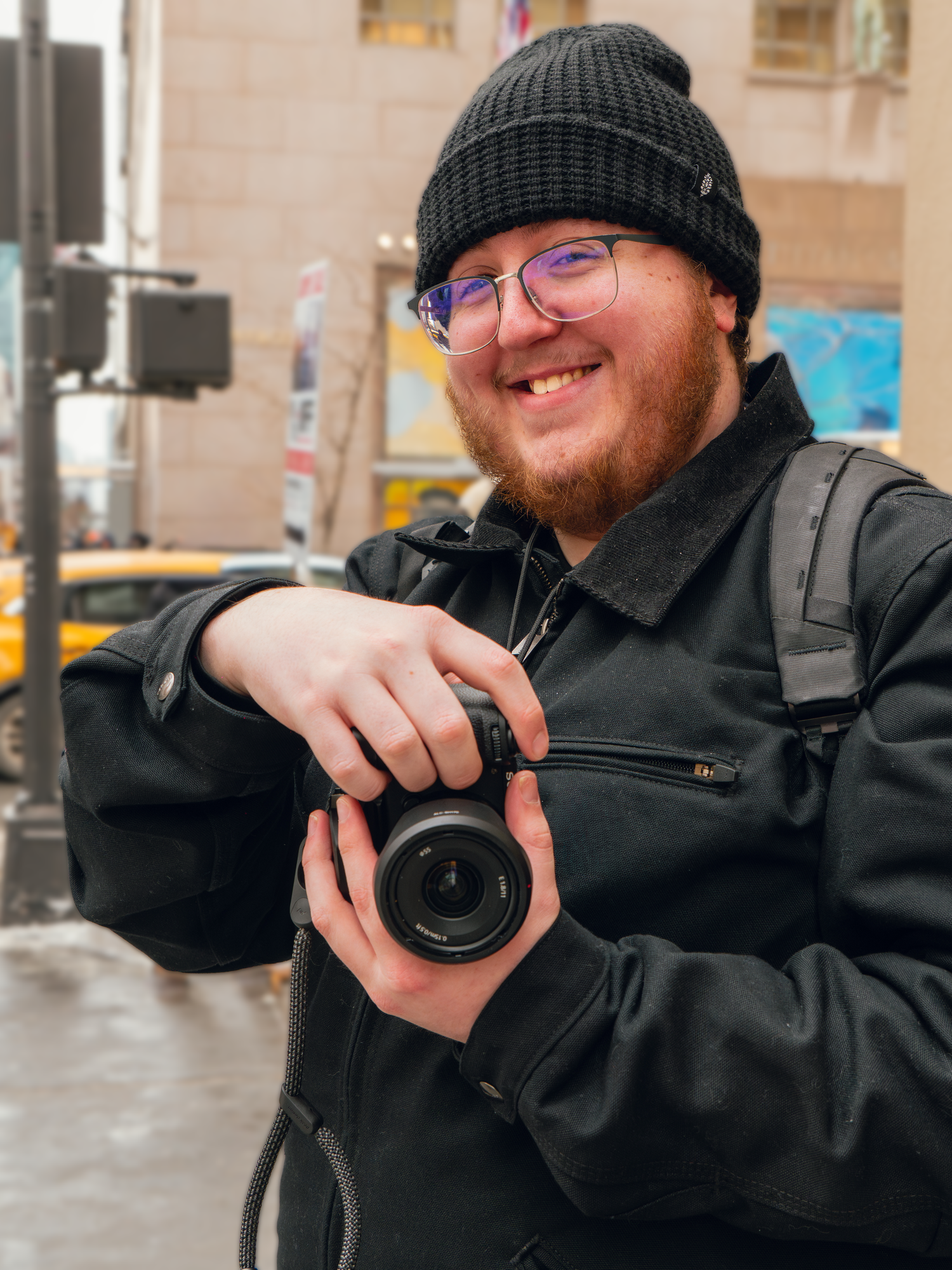 A cheerful bearded photographer in a black beanie and glasses holding a camera on a city street.