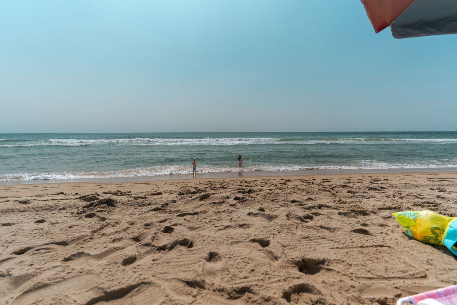 Sandy beach with gentle ocean waves, two people standing near the shoreline under a clear blue sky on a sunny day.