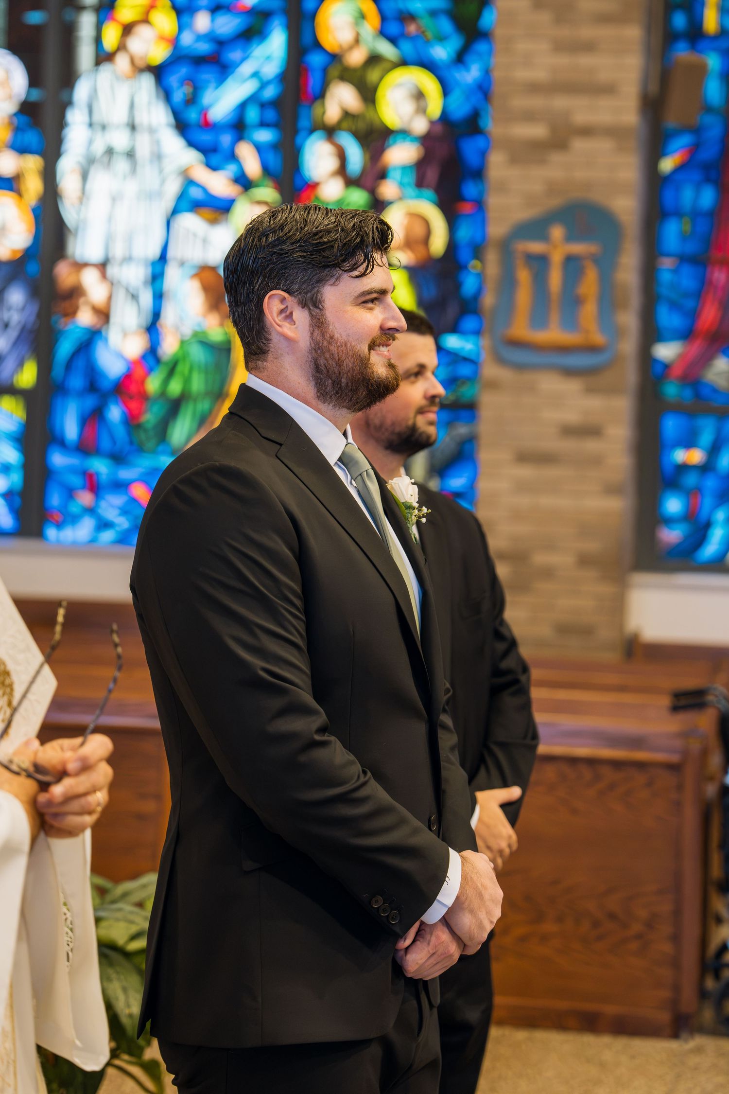 Groom smiling at altar in dark suit with boutonniere, stained glass windows behind him in church.