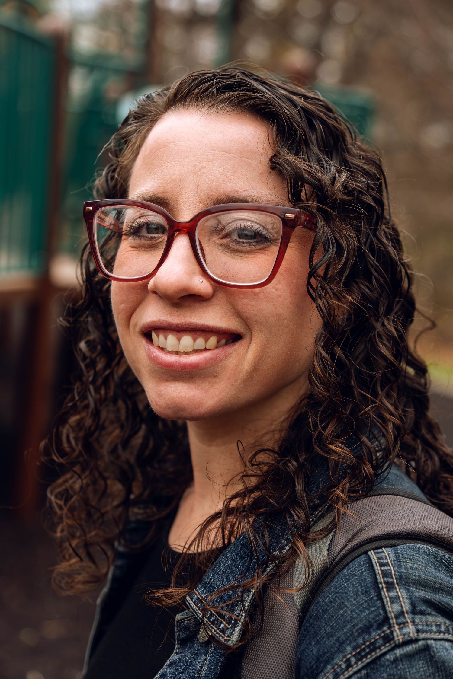 Smiling woman with curly hair and red glasses wearing a denim jacket outdoors.