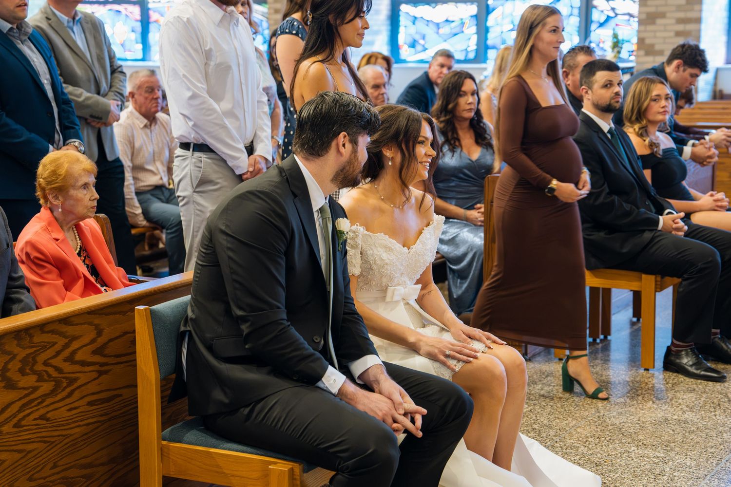 Bride and groom sit together in church pew during wedding ceremony with guests standing in background.
