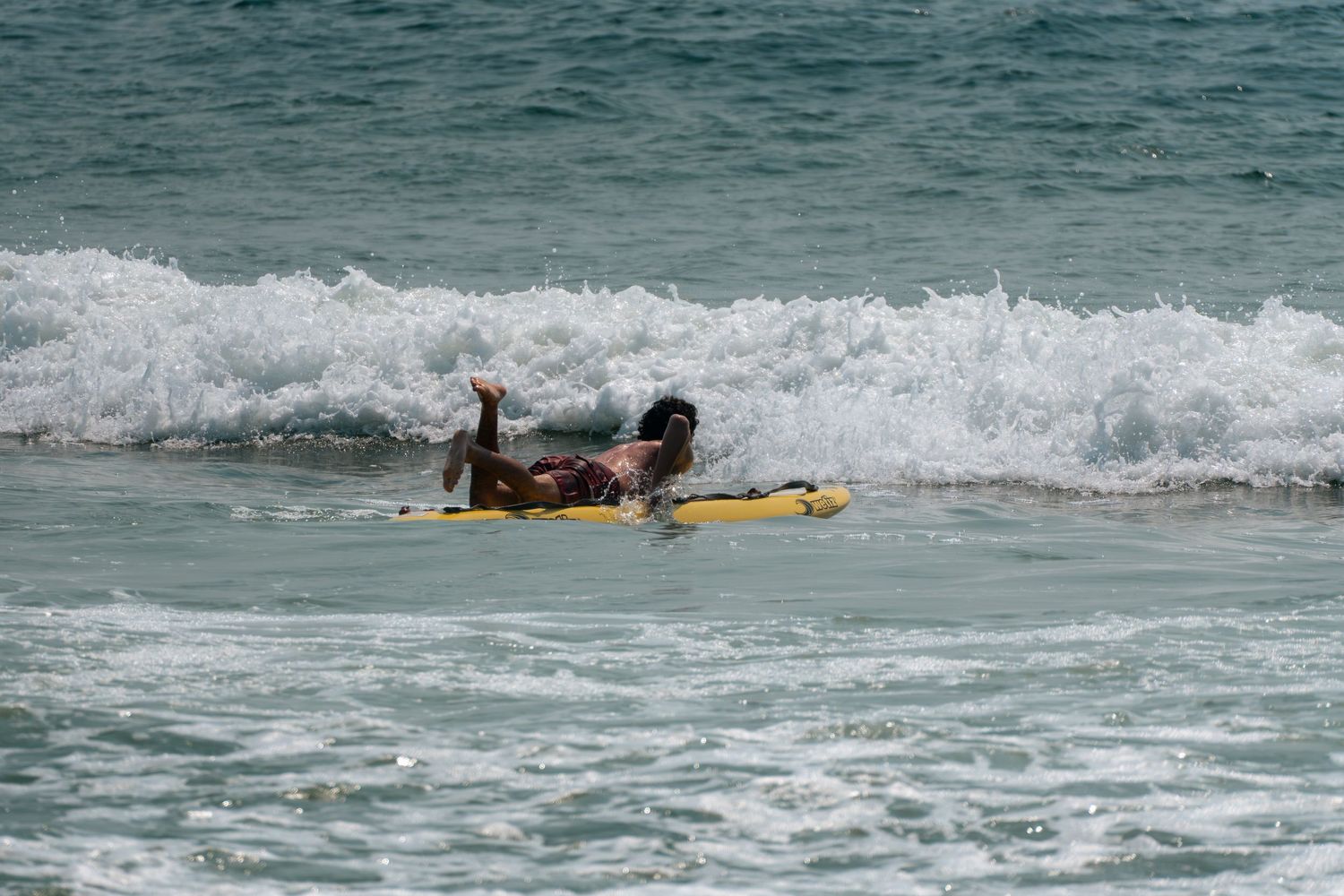 Person lying on a yellow surfboard riding ocean waves at the beach.