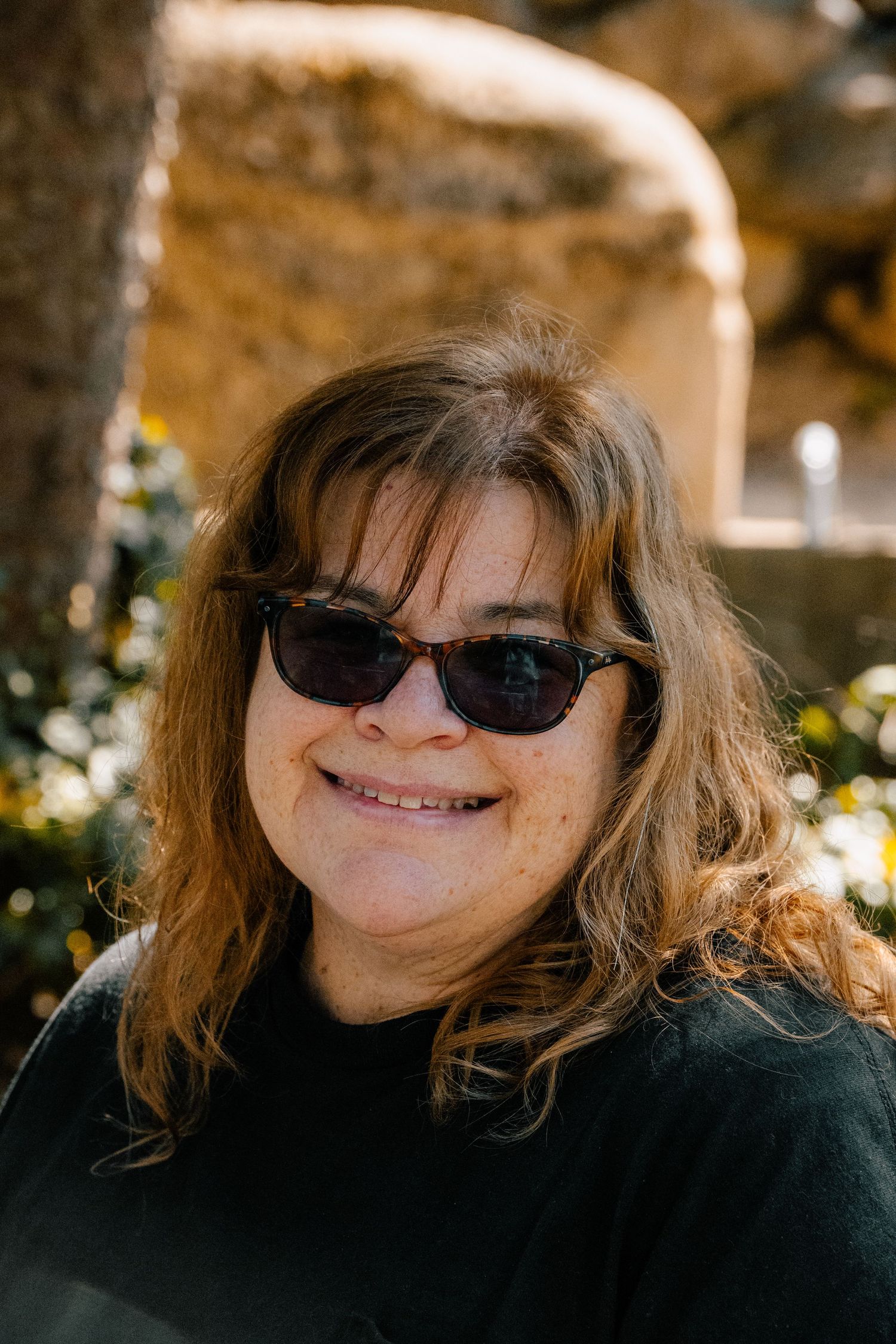A smiling middle-aged woman with wavy auburn hair wearing dark sunglasses and a black top outdoors in natural sunlight.