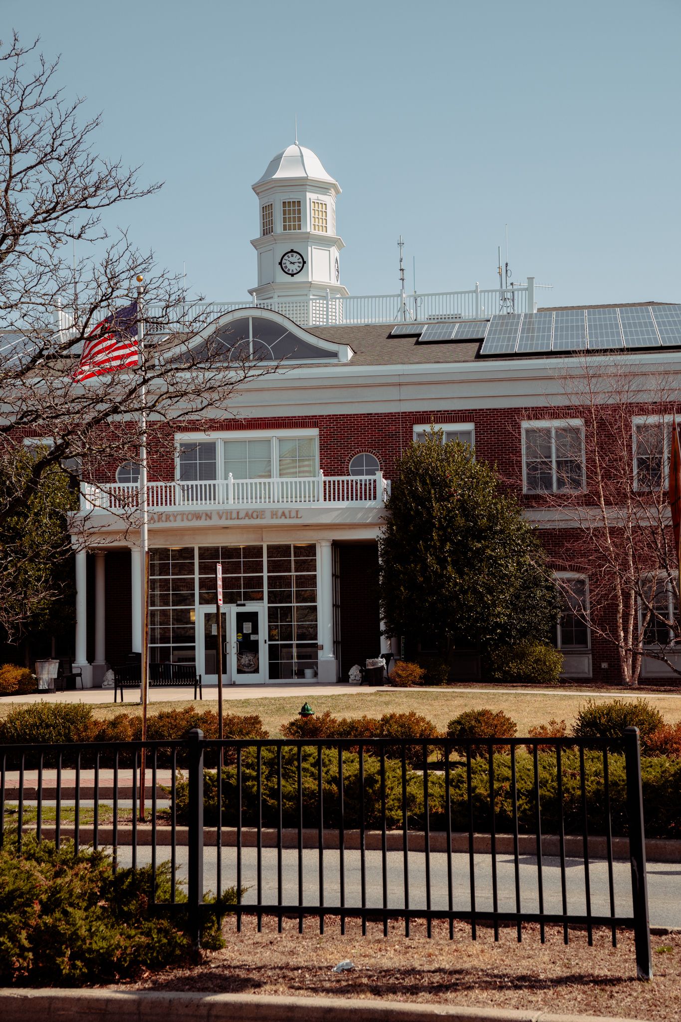 Historic red brick Levittown Village Hall with white clock tower, American flag, and solar panels on a clear day.