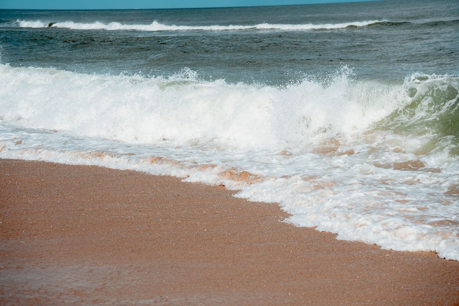 Powerful white-capped ocean wave crashing onto a reddish-brown sandy beach shoreline.