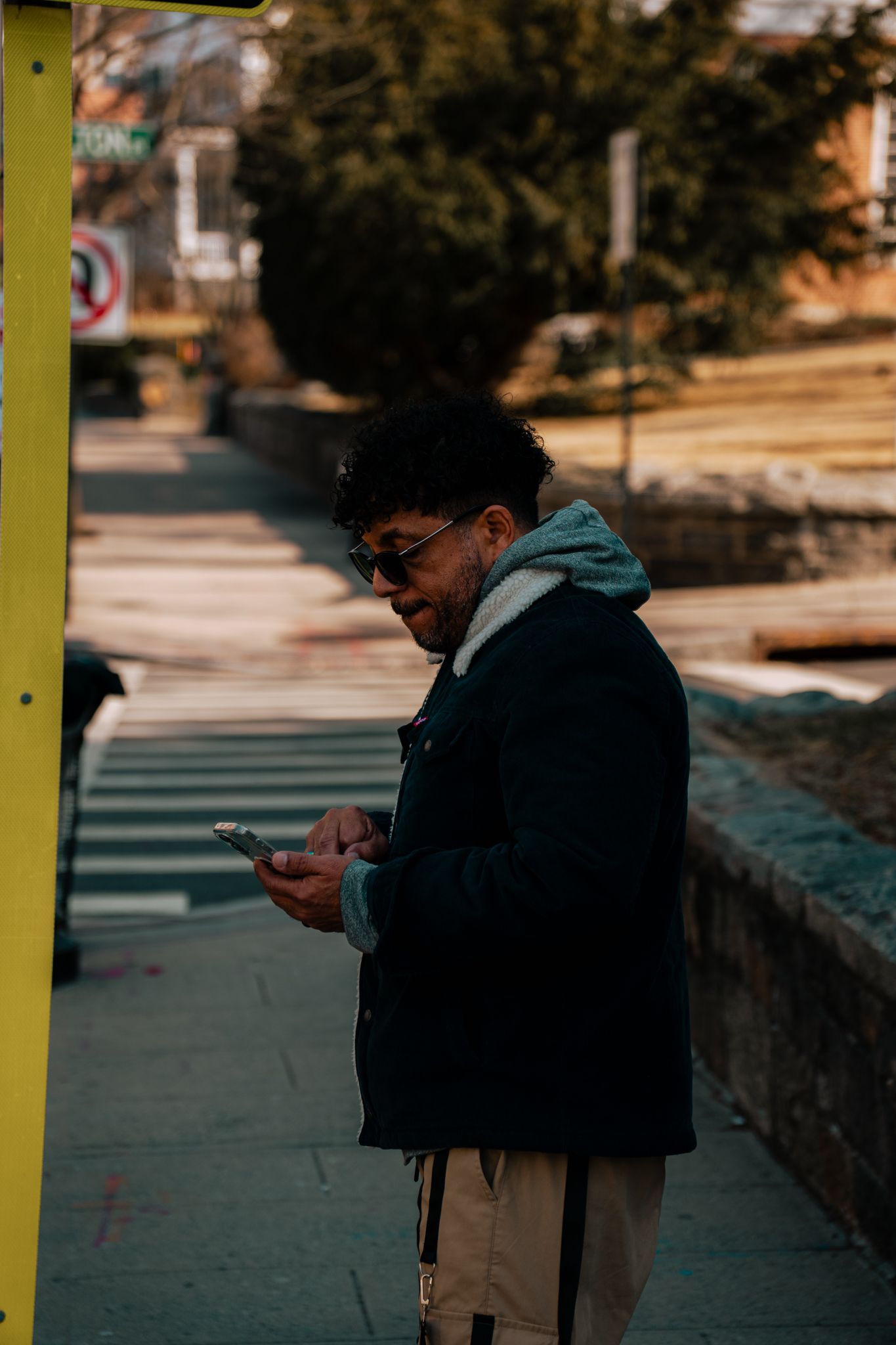 Young person wearing glasses and dark puffer jacket browsing smartphone on city sidewalk.