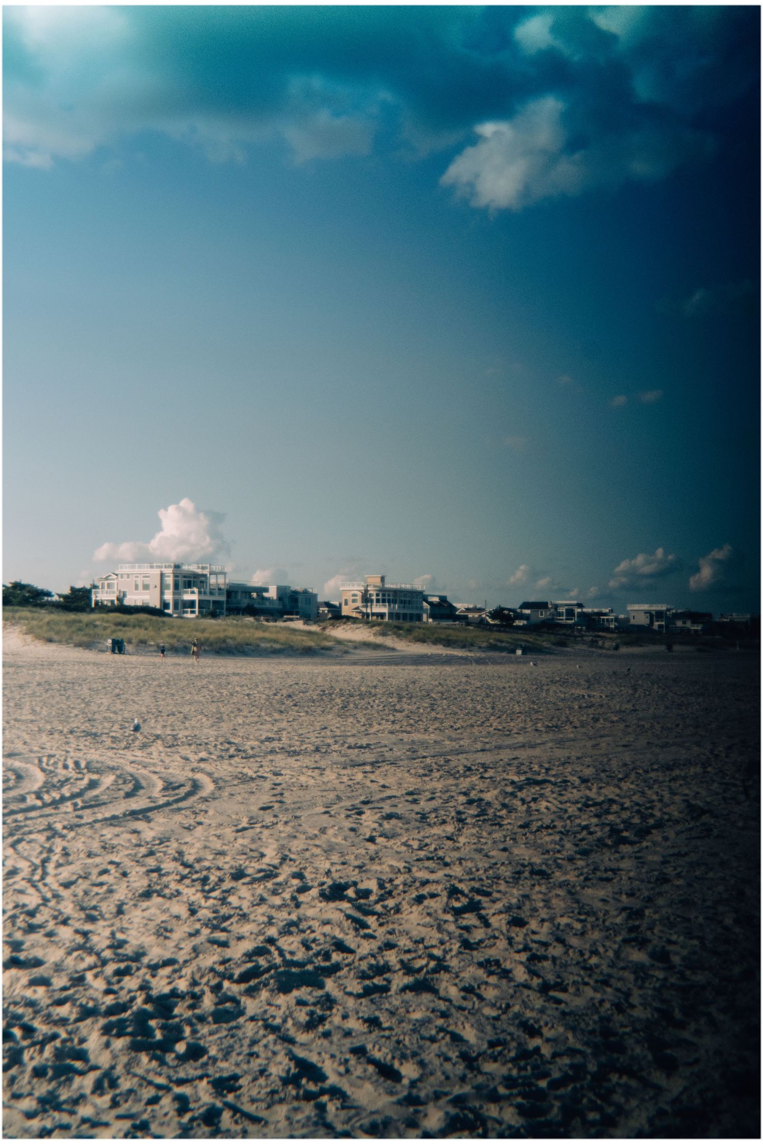Wide sandy beach stretching toward beachfront homes under a dramatic blue cloudy sky.