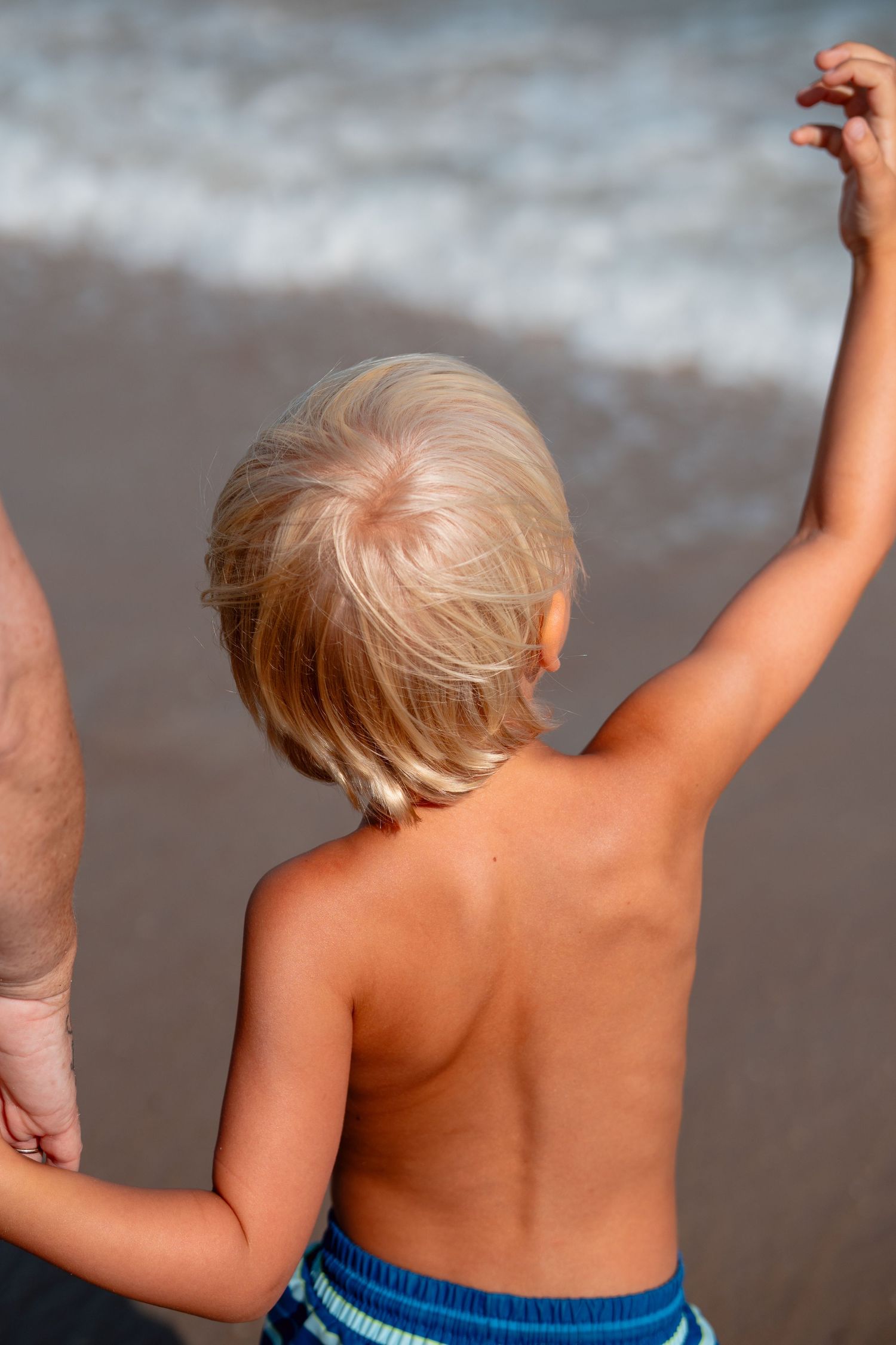 Blonde toddler in striped swim trunks holding an adult hand while raising arm toward ocean waves.