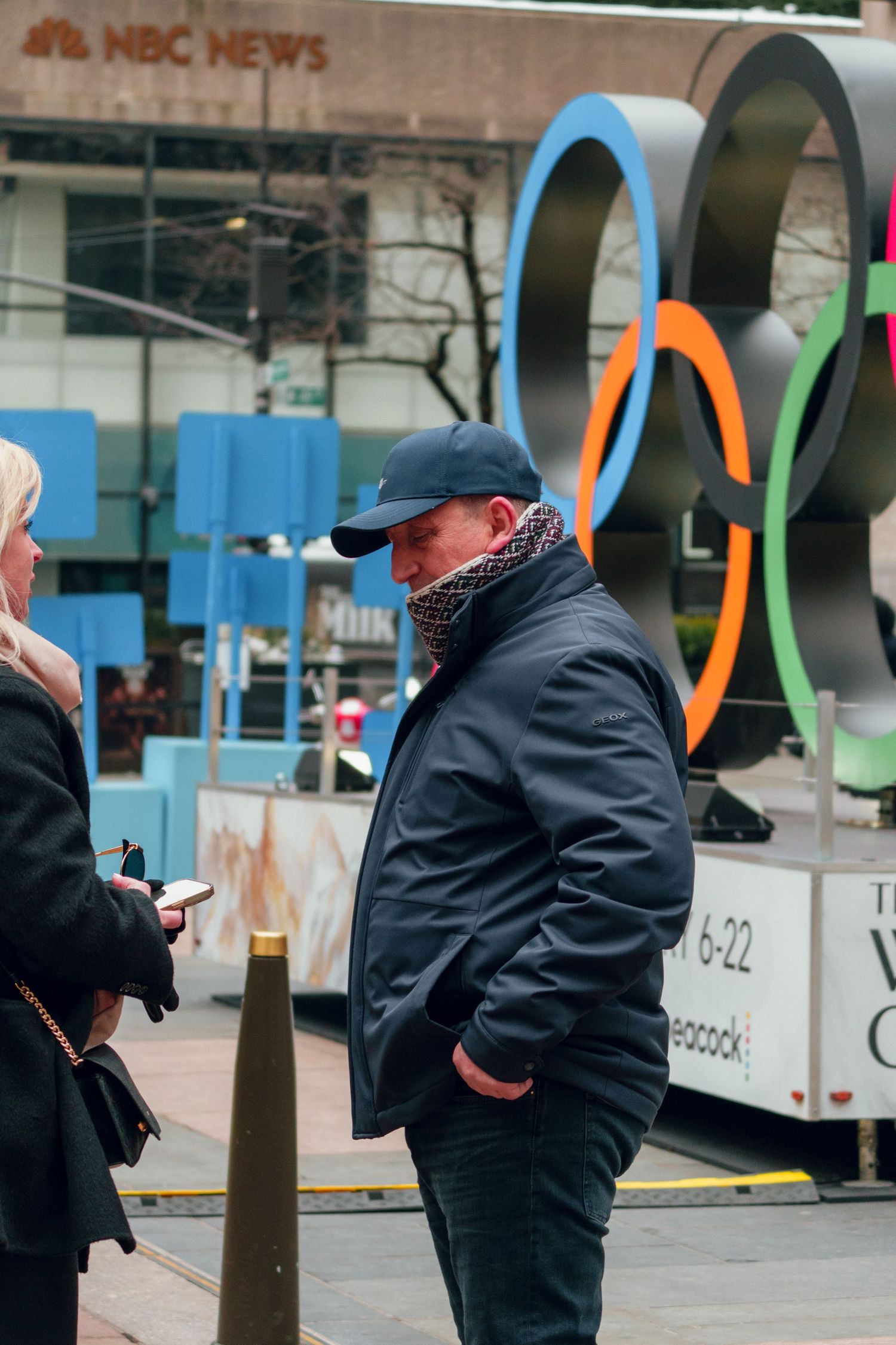 Man in blue cap and scarf standing near NBC News studio with Olympic rings display in NYC.