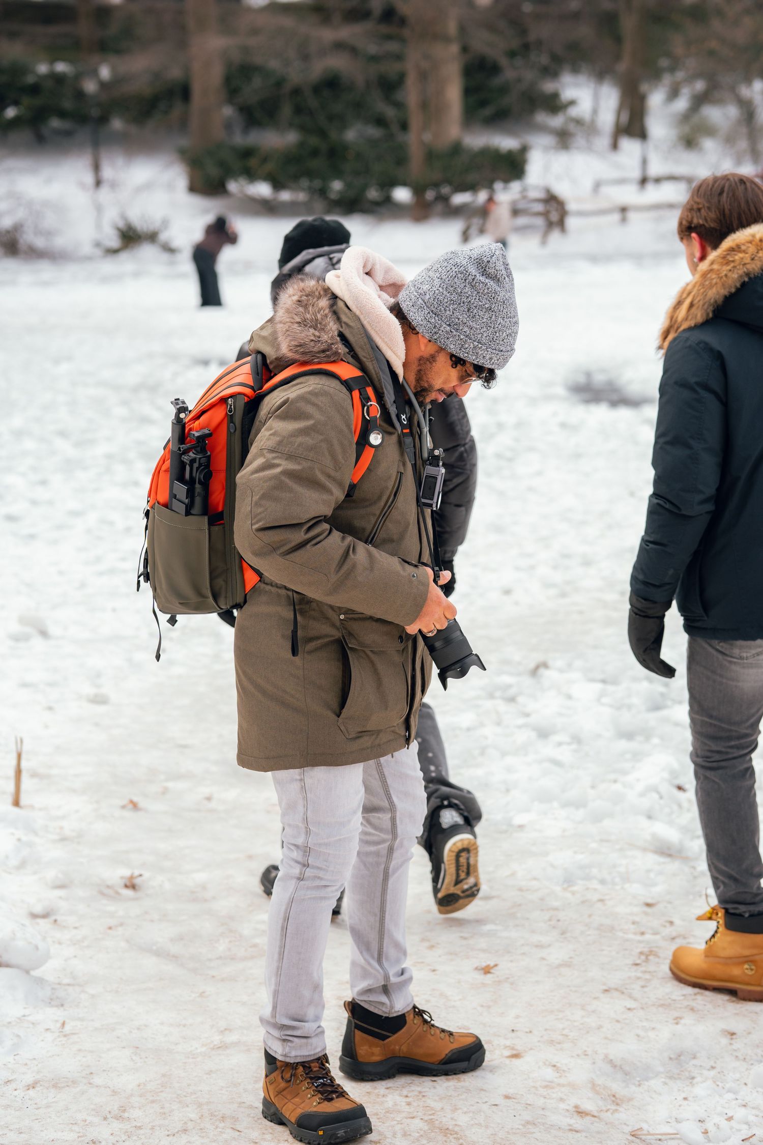 Male photographer with orange backpack checking camera gear in a snowy winter park setting.