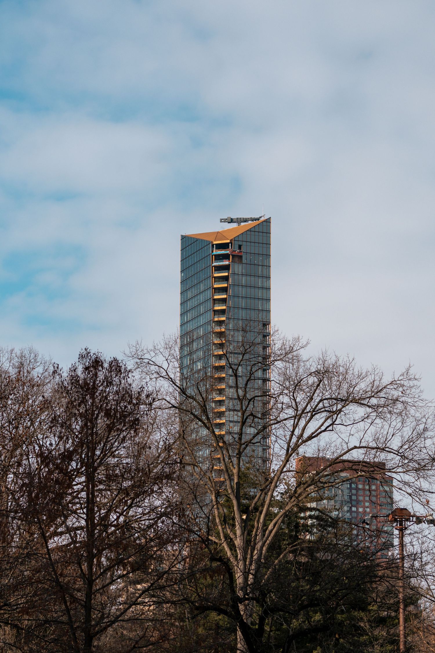 Tall glass skyscraper with golden angular top rises above bare winter trees under cloudy sky.