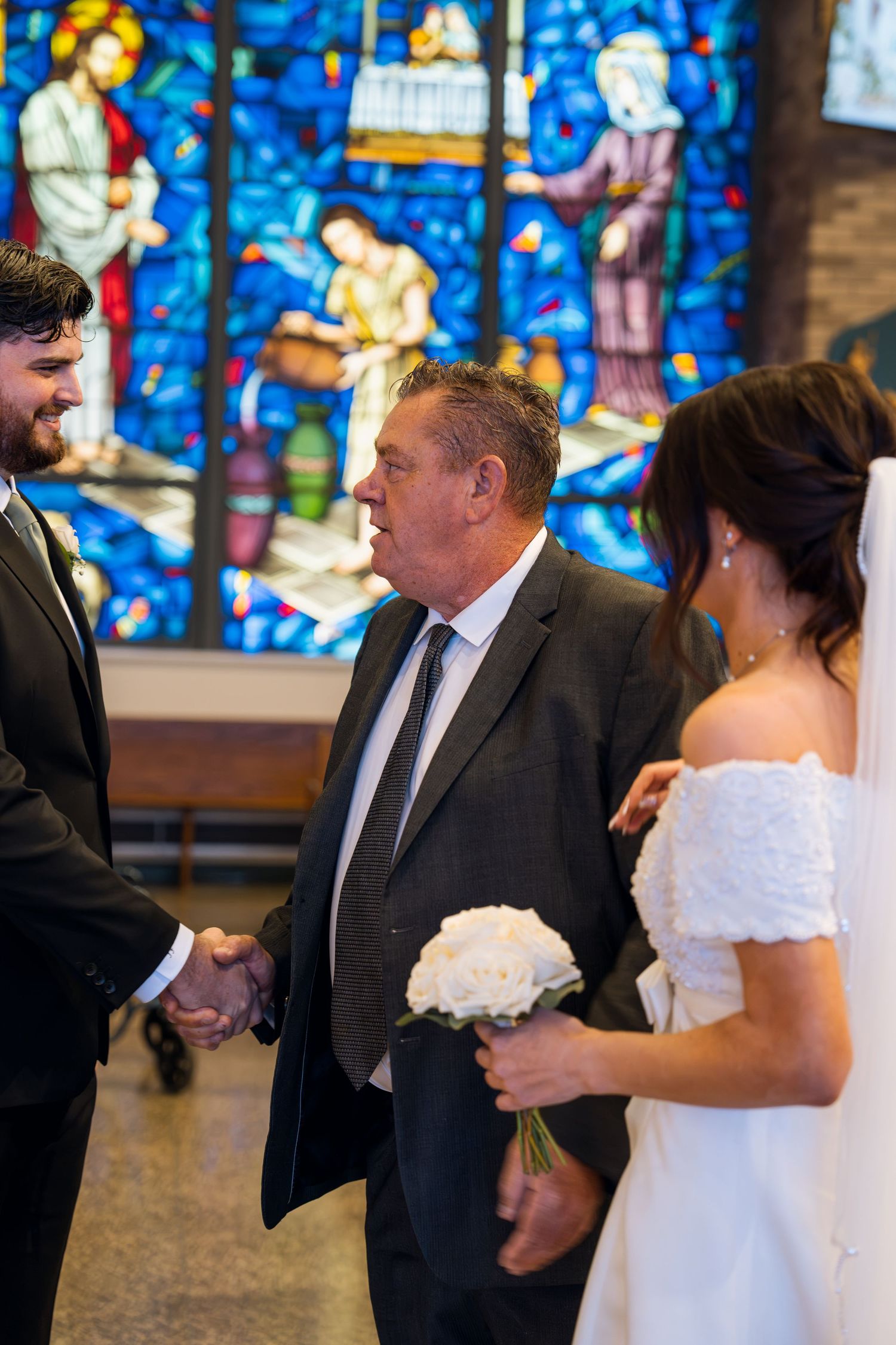 Father of the bride shaking hands with groom at church wedding ceremony, bride holding white bouquet near stained glass.