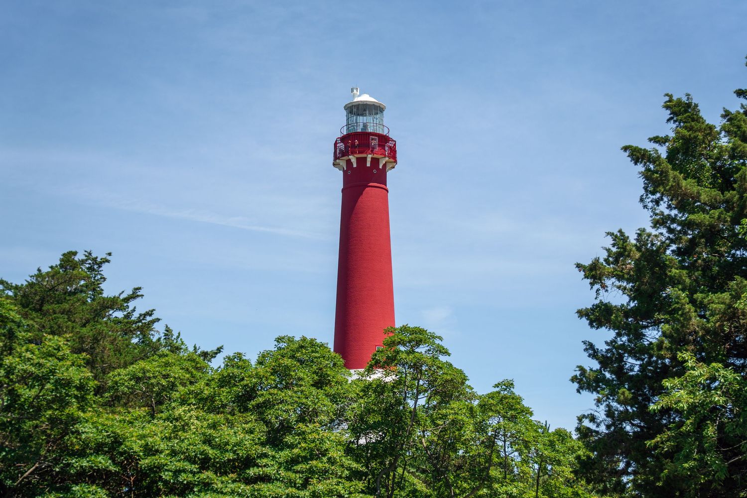 Tall red lighthouse rising above lush green trees against a clear blue sky.