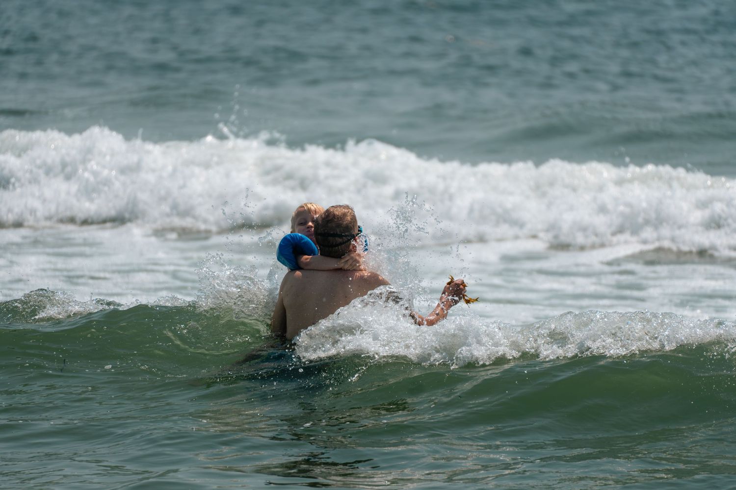 Adult carrying a child through ocean waves at the beach, navigating rough surf together.