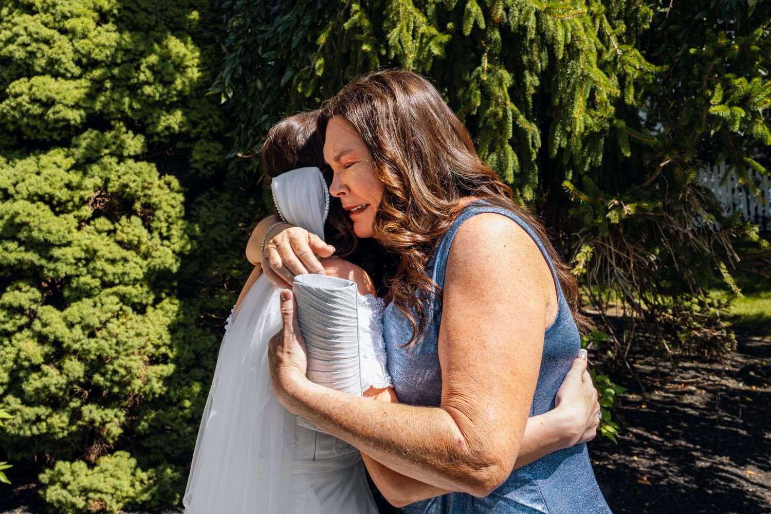 A bride in a white dress shares an emotional hug with a woman in a blue dress outdoors near green trees.