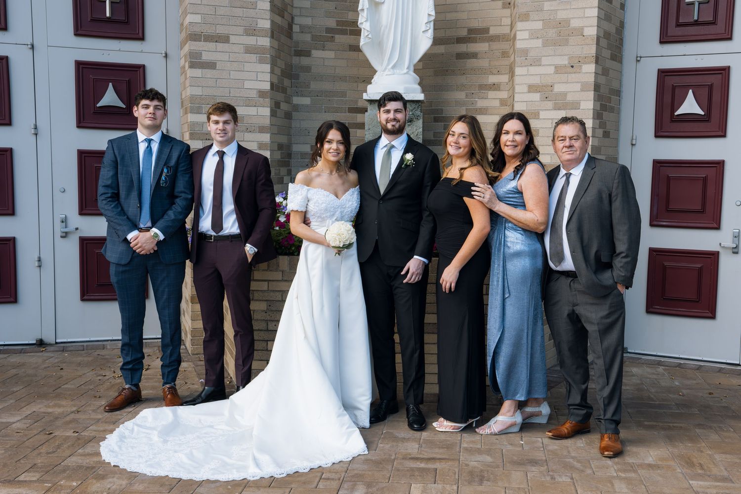 Wedding party poses outside a brick church, bride in white gown with groom and family in formal attire.