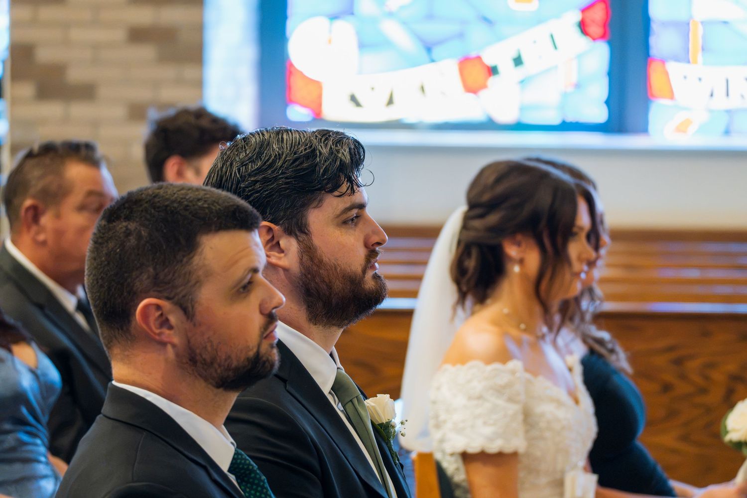 Guests seated in church pews watch a bride in a lace off-shoulder gown during a wedding ceremony.