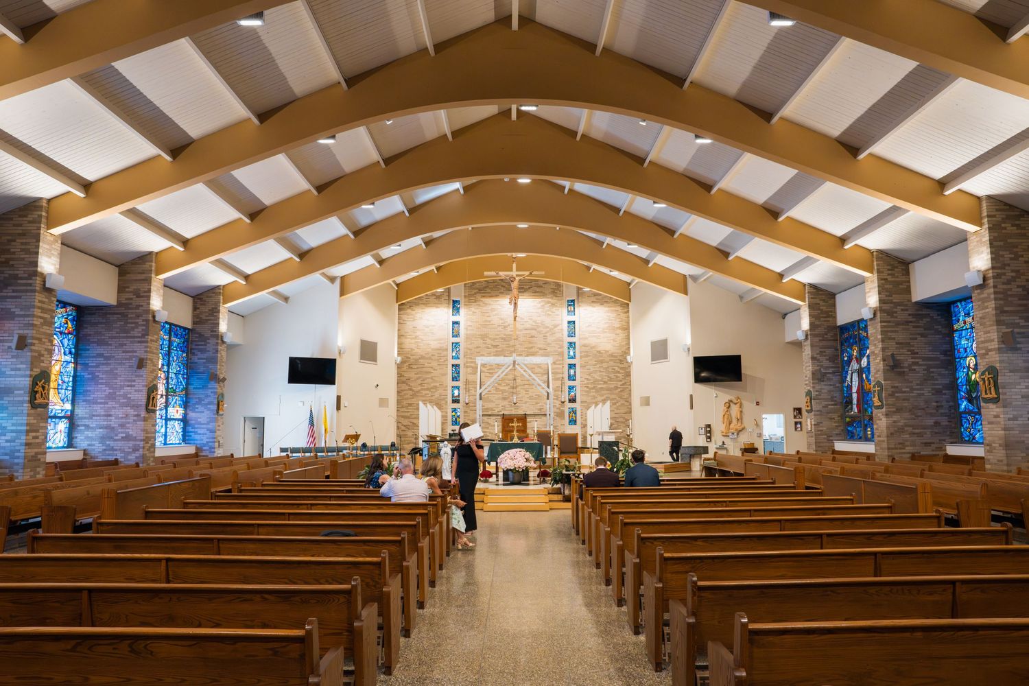 Bright church interior with wooden pews, arched ceiling beams, stained glass windows, and altar at the front.