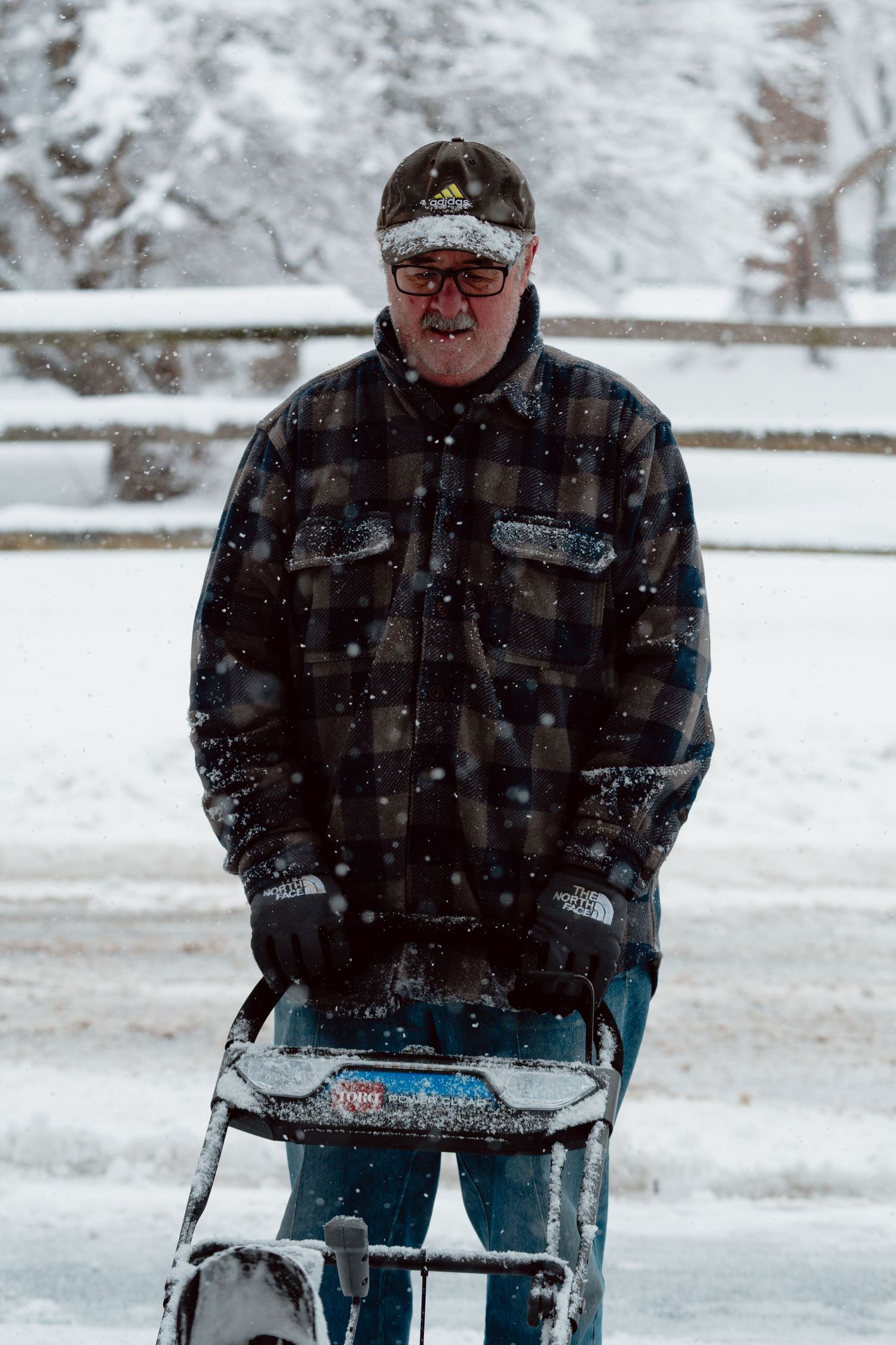 Older man in plaid flannel jacket pushing a snowblower through heavy falling snow outdoors.