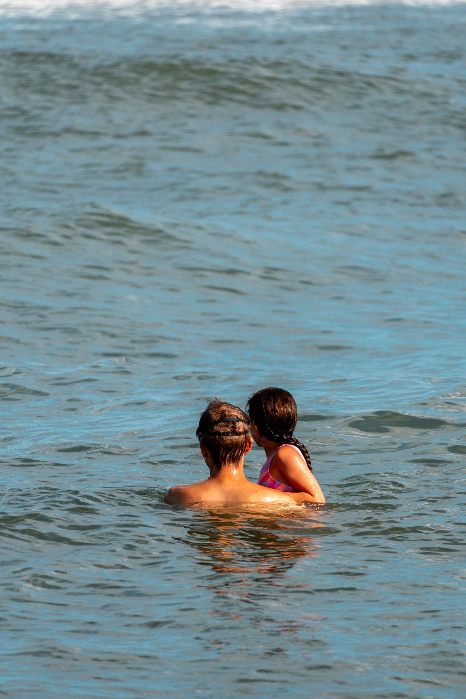 Two swimmers seen from behind in calm blue ocean water near the shore.