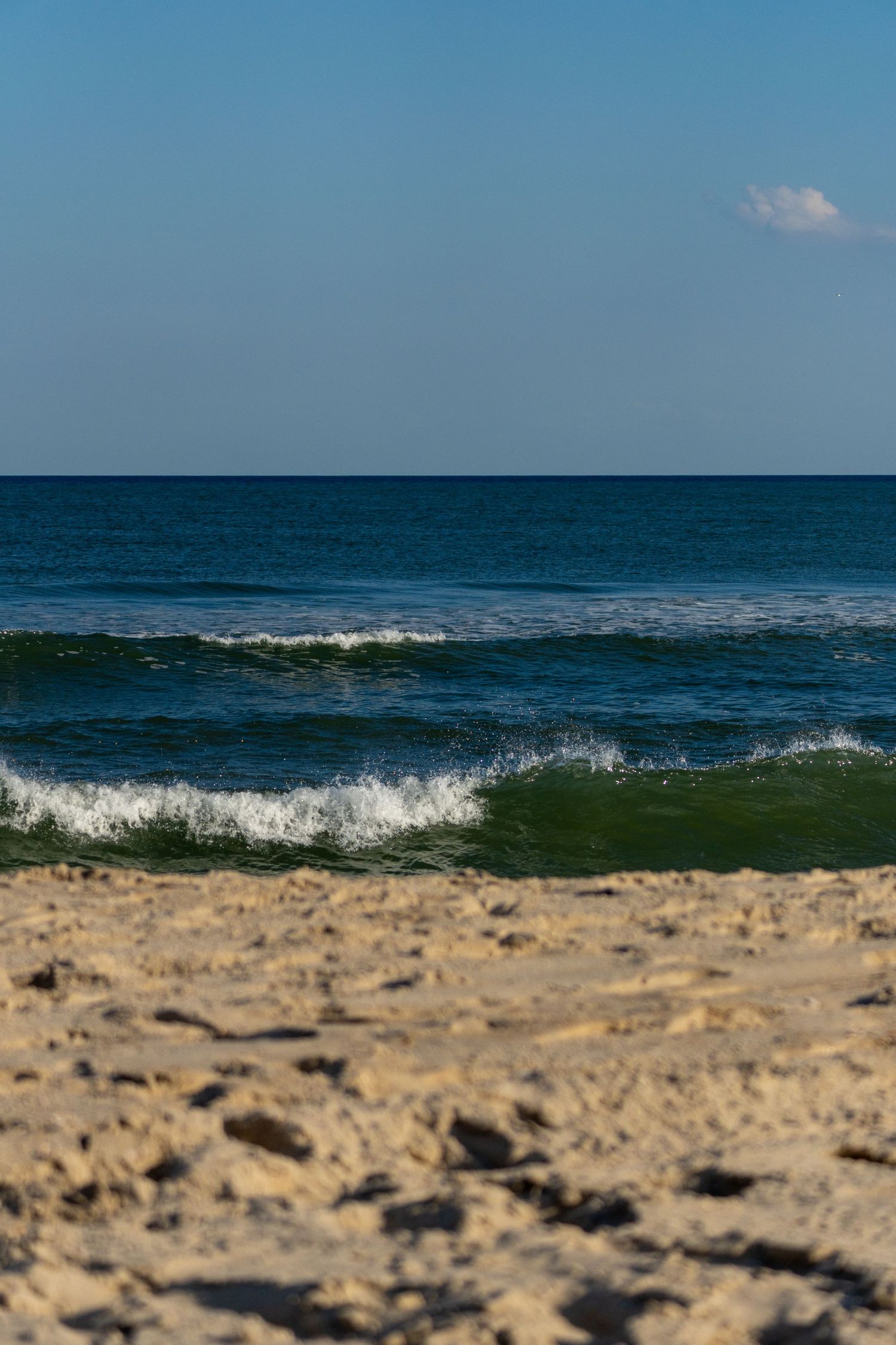 A crashing ocean wave rolls onto a sandy beach with deep blue water and a partly cloudy sky above.