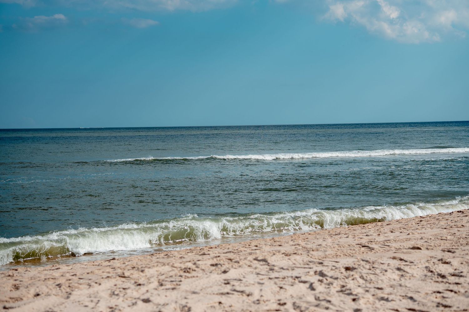 Gentle waves washing onto a sandy beach with a partly cloudy blue sky horizon.