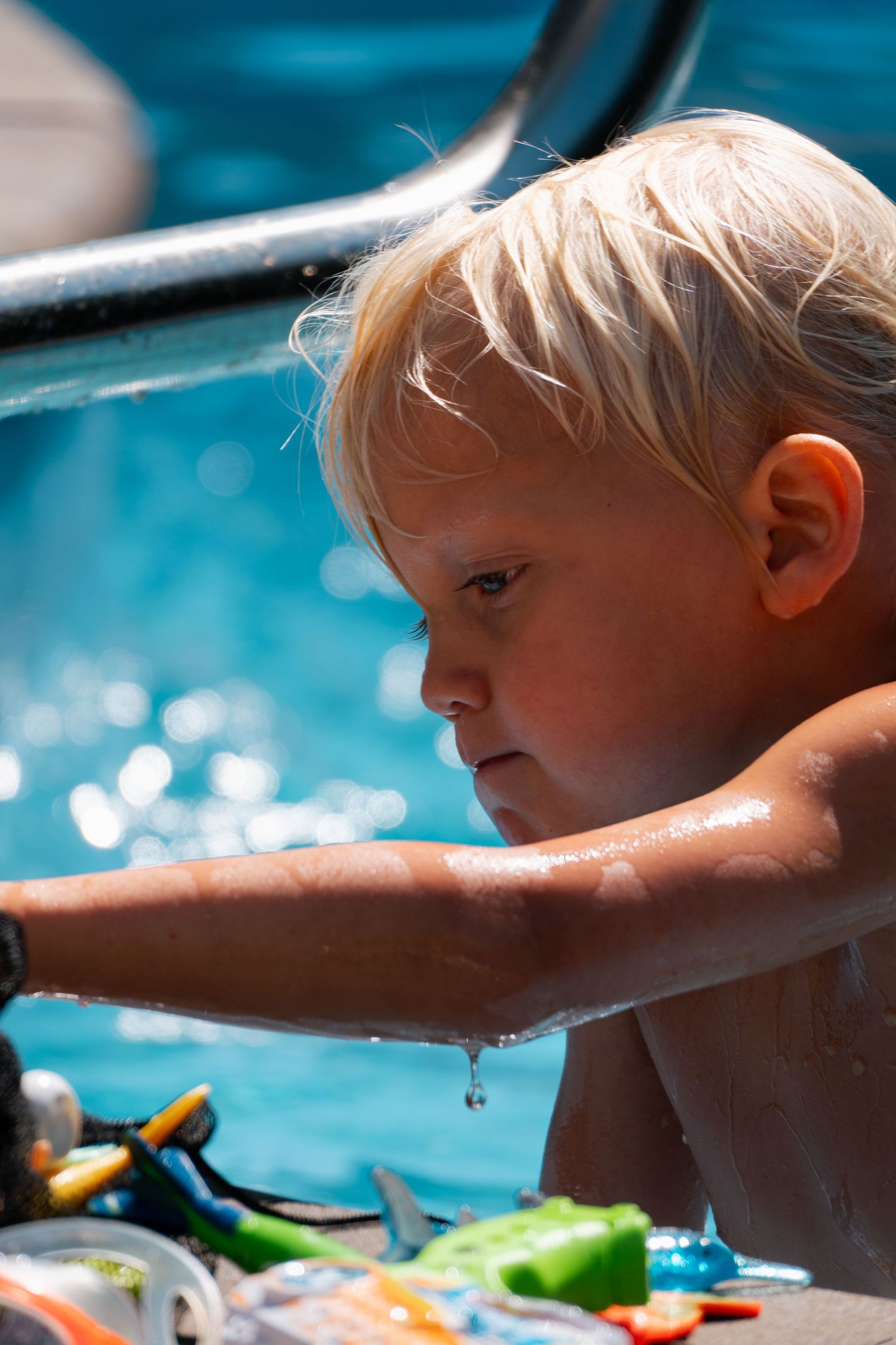 Blonde toddler boy reaching over pool edge with water dripping from his hand near toys.