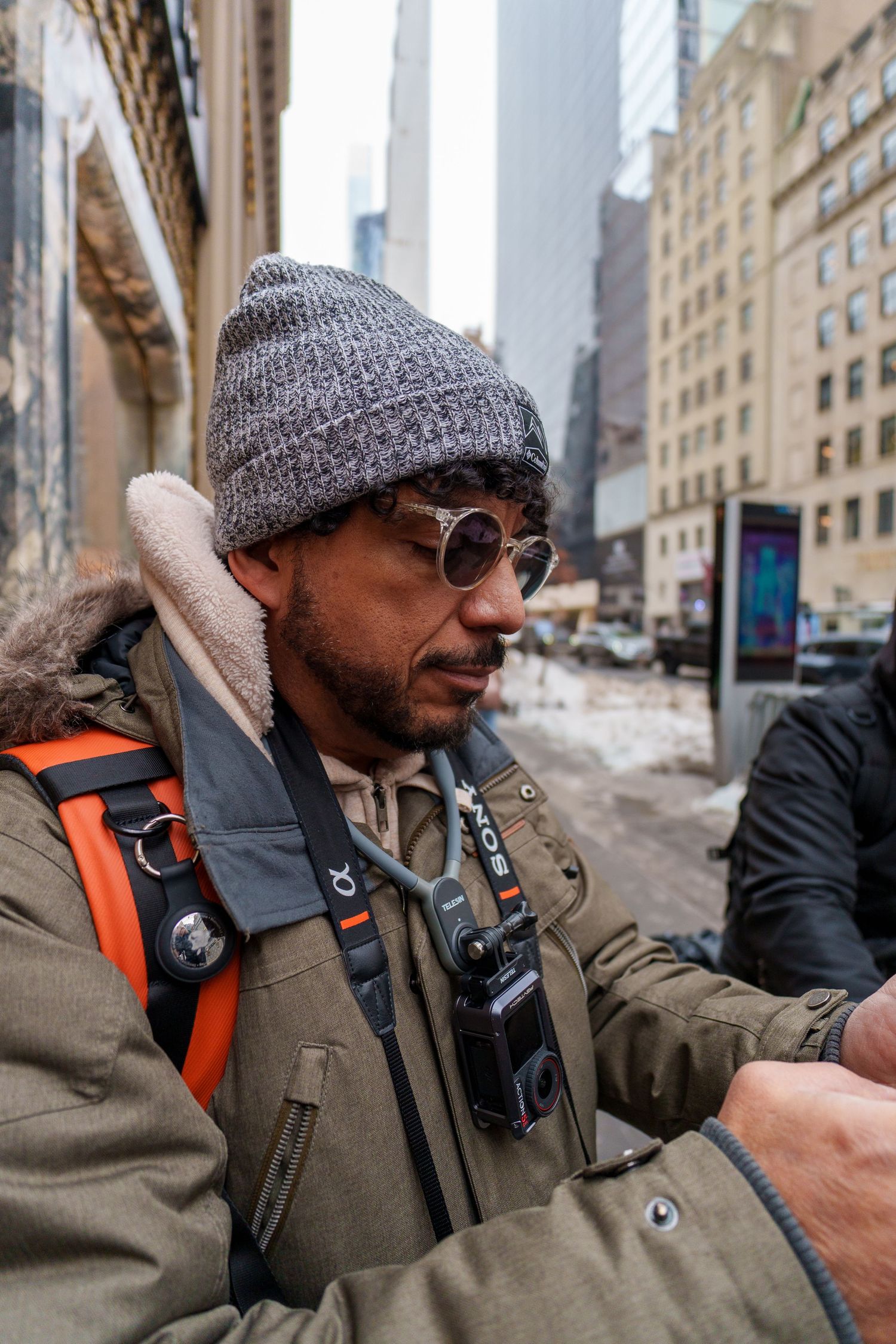 Close-up of a man in a gray beanie and round sunglasses on a snowy urban street in New York City.