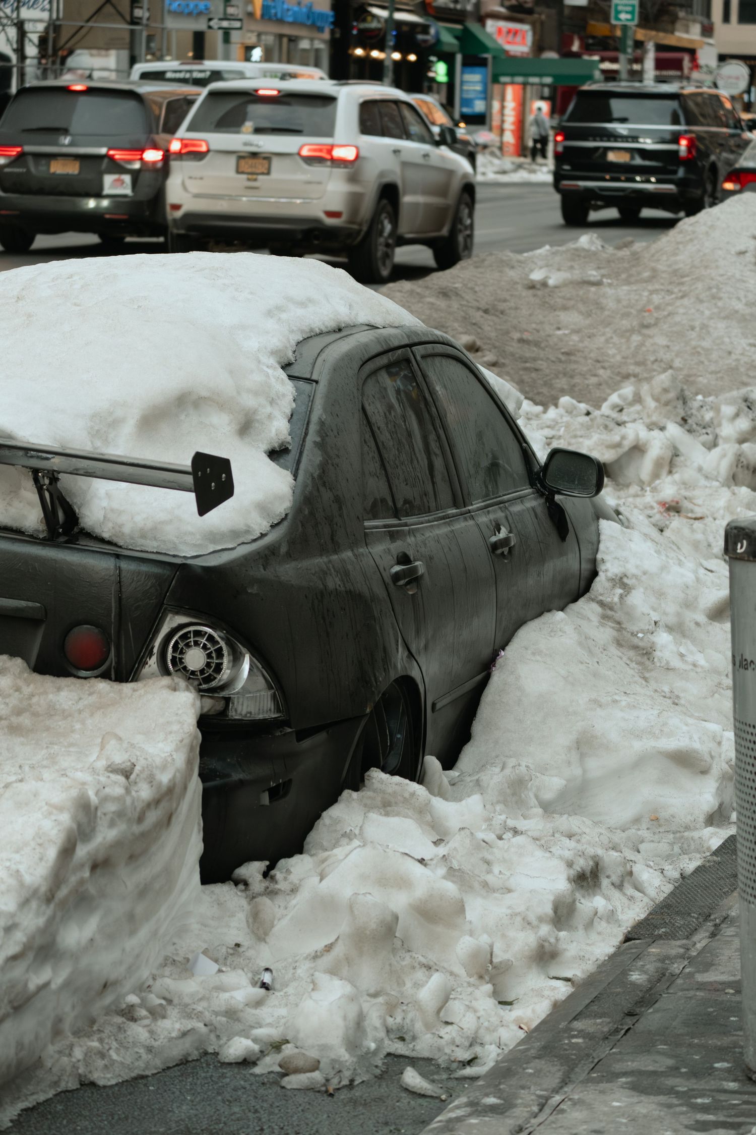 A snow-covered parked car is buried in a snowbank along a busy New York City street.