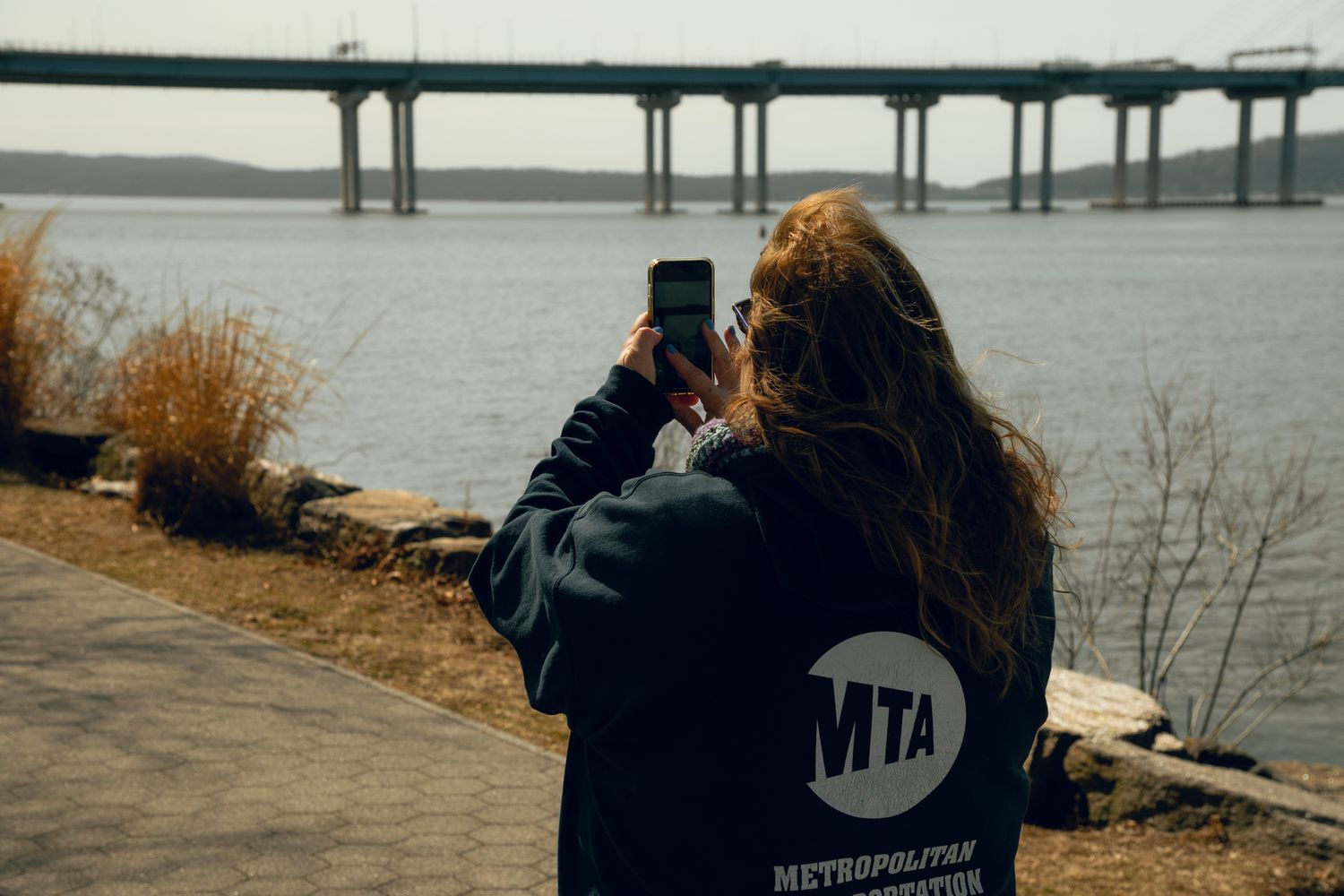 Woman in MTA Metropolitan Transportation jacket photographs a cable-stayed bridge over a wide river.