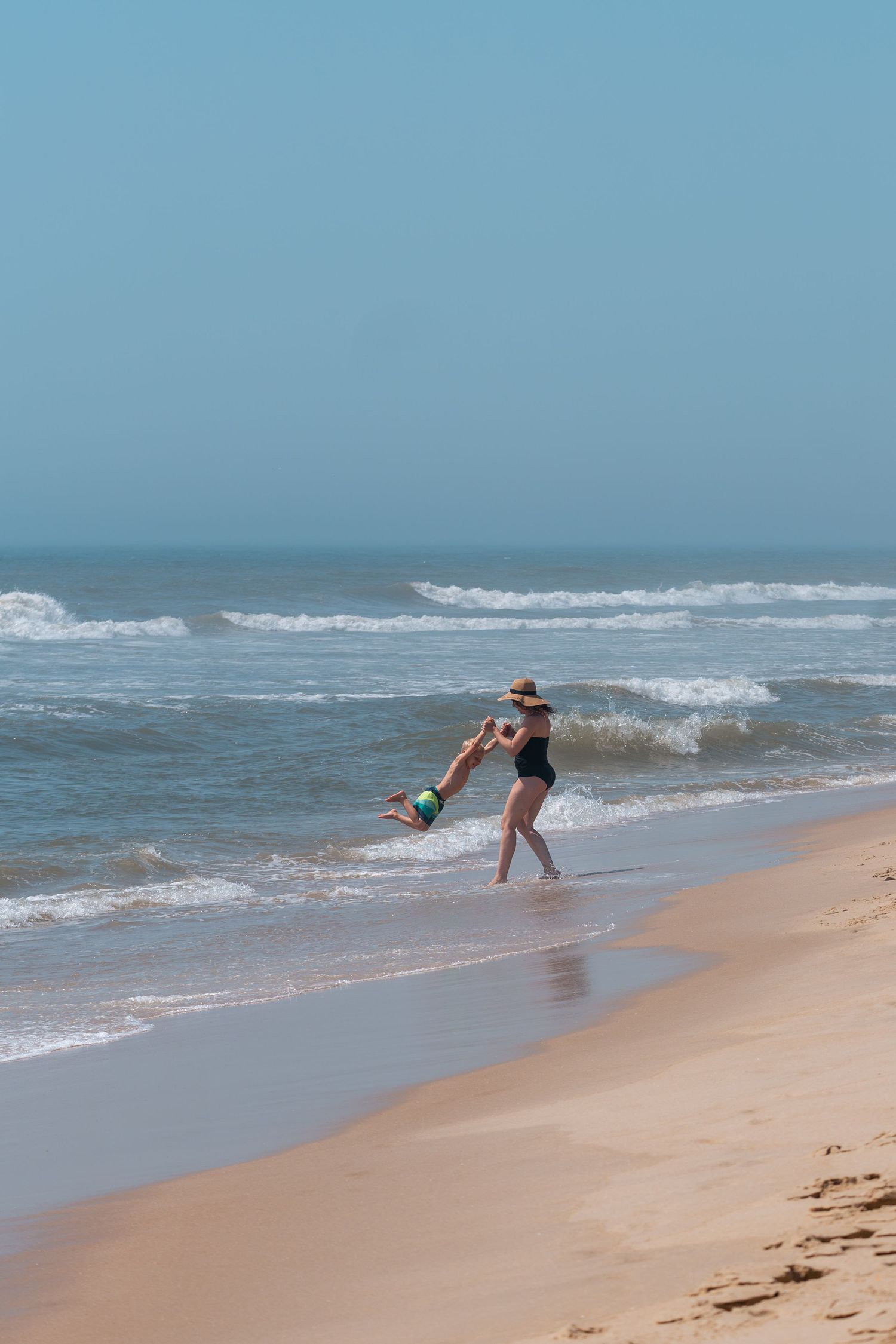 Woman in black swimsuit and hat swinging a child on a sunny sandy beach with ocean waves in the background.