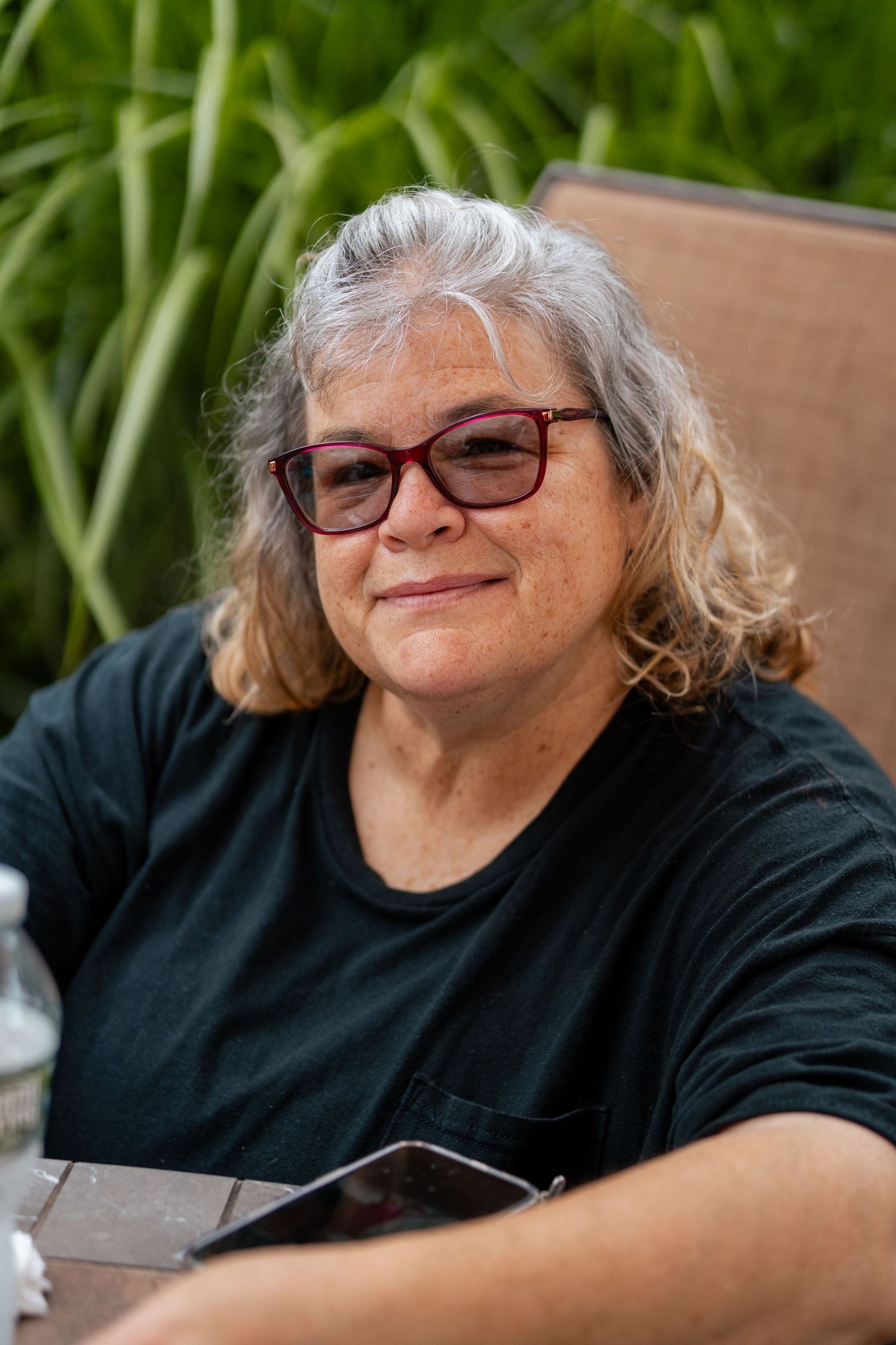 Smiling older woman with gray hair and red glasses sitting outdoors near green plants.