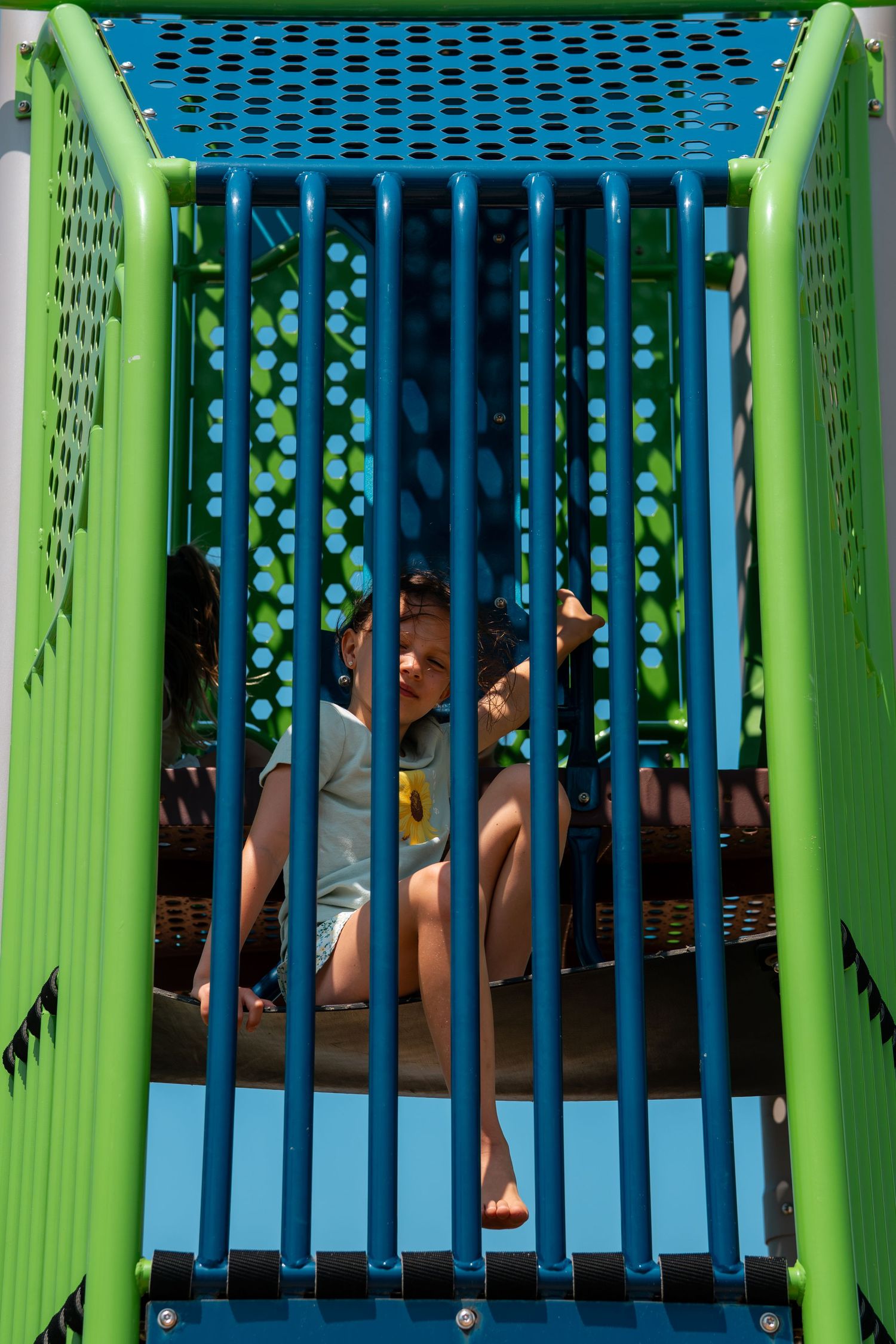 Children playing inside a colorful blue and green playground structure on a sunny day.