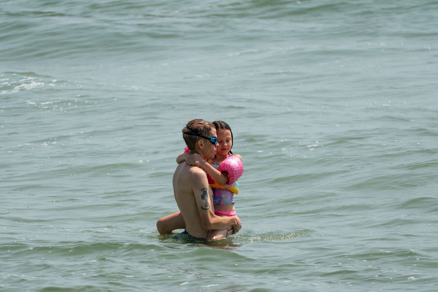 Adult holding a child in shallow ocean water at the beach on a sunny day.