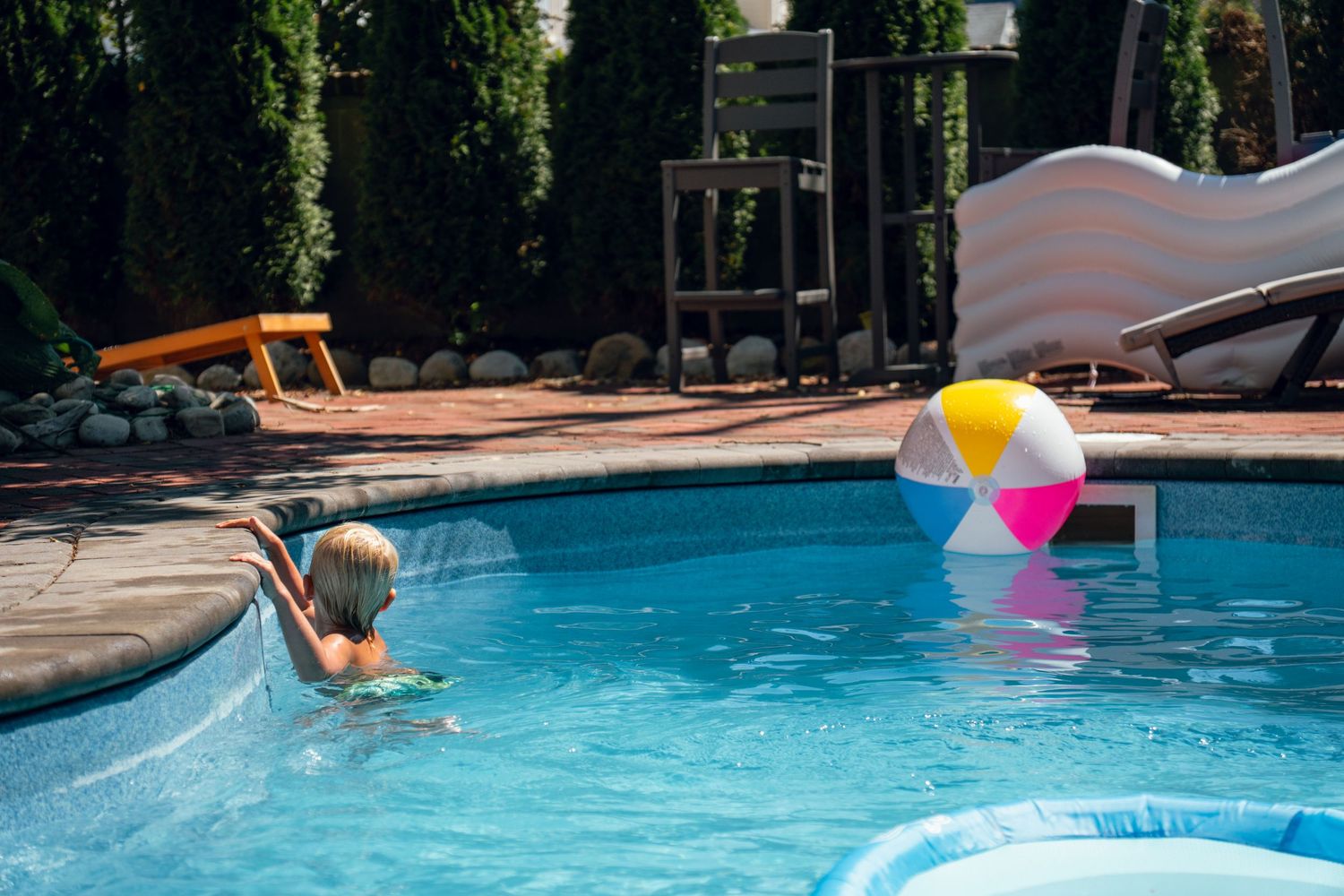 Blonde child reaching for a colorful beach ball floating in a sunny backyard swimming pool.