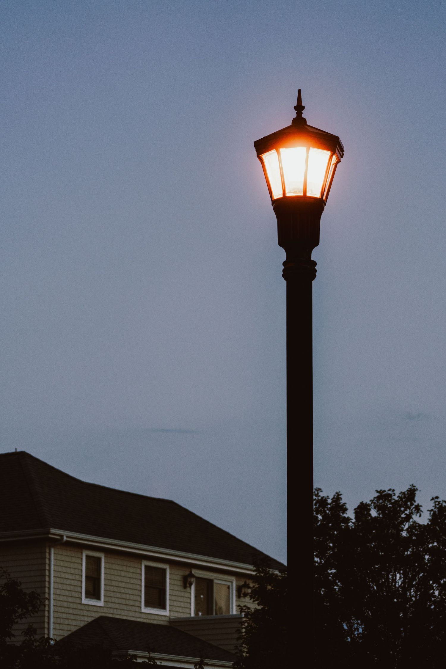 Ornate black street lamp glowing warmly against a twilight blue sky near a residential home.