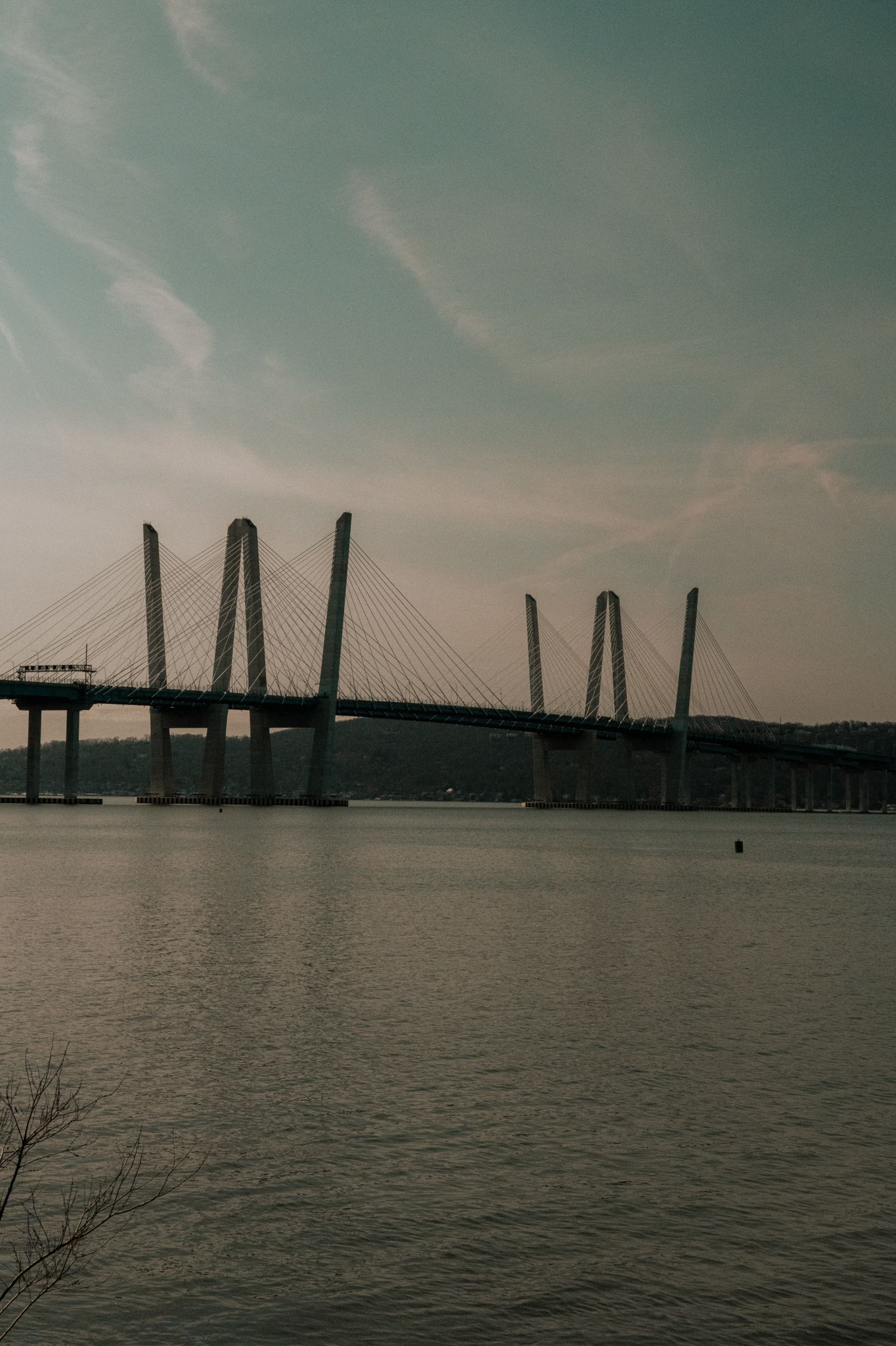 Cable-stayed bridge with multiple tall towers reflects over calm river water under a teal-toned sky.