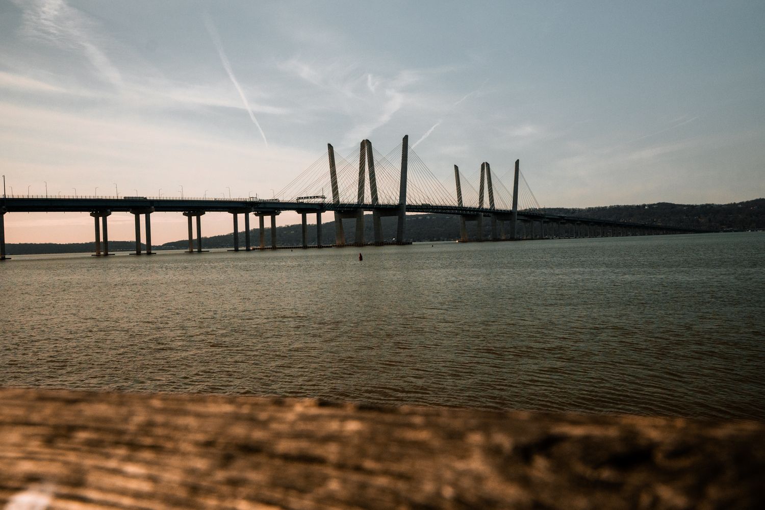 A cable-stayed bridge stretches across a calm river at dusk, viewed from a wooden dock foreground.