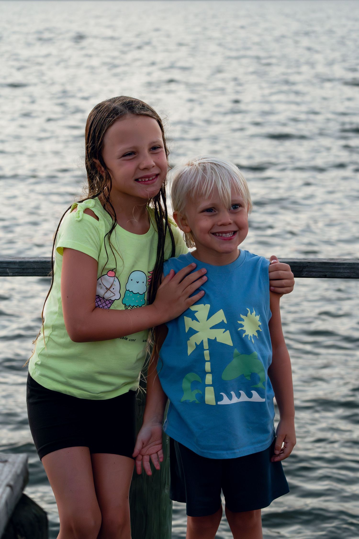 Two smiling children, a girl and boy, posing together on a dock with water in the background.