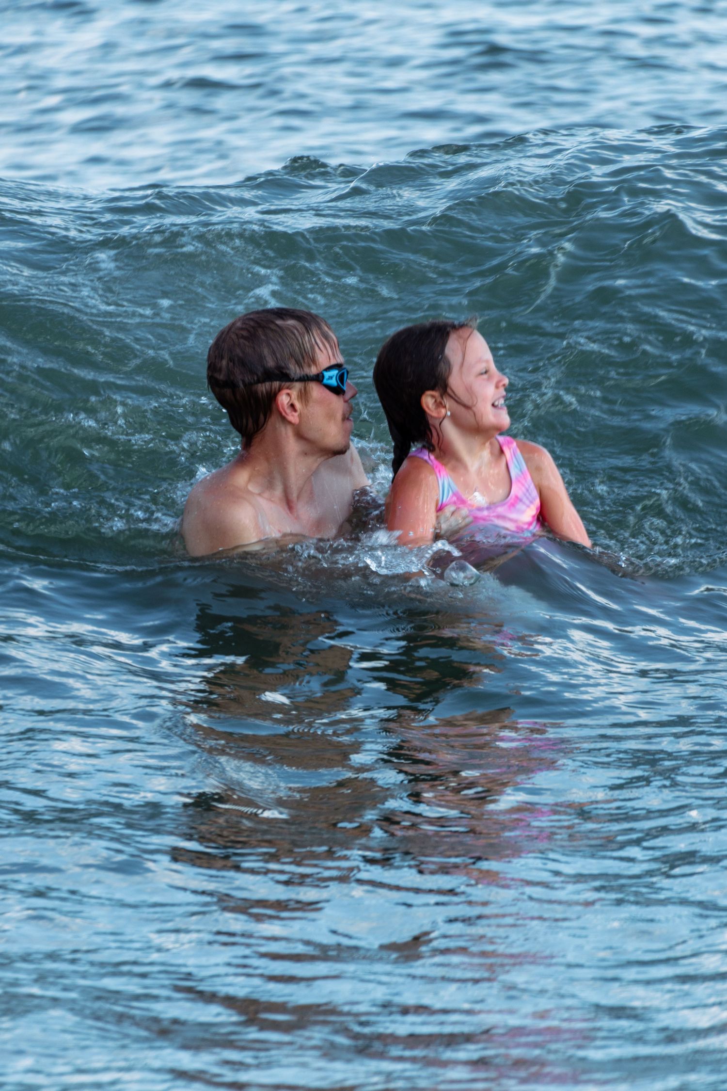 A man in blue goggles helps a young girl in a pink swimsuit swim in ocean waves.